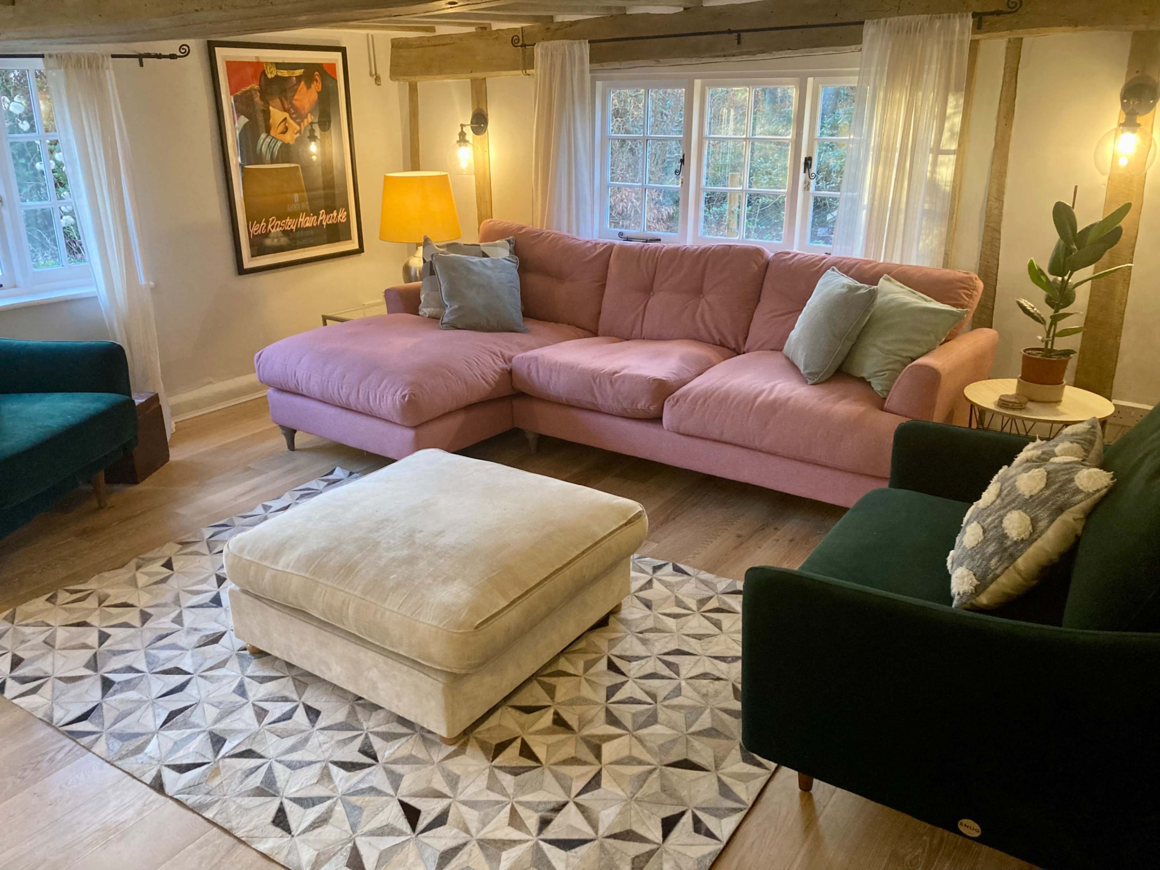 A cozy living room with a pink sectional sofa, a green armchair, a light-colored ottoman on a patterned rug, and large windows allowing natural light to enter.