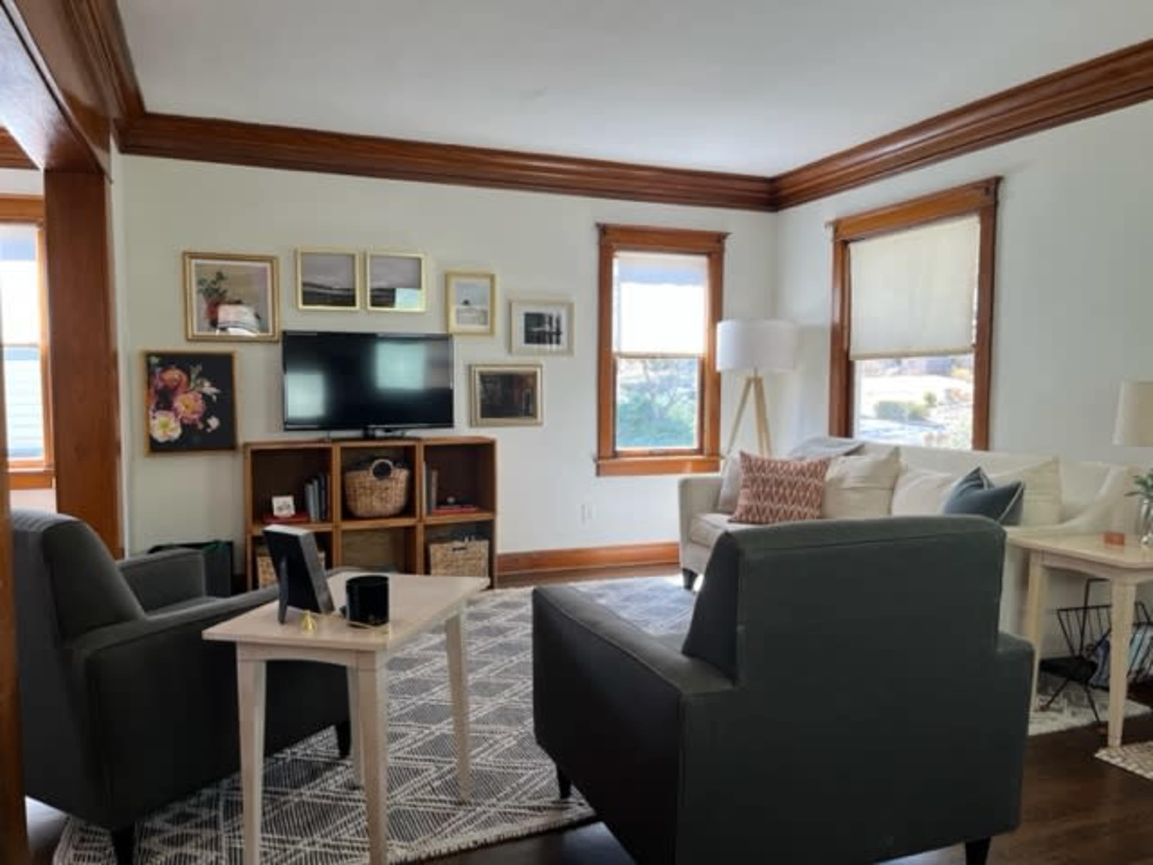A cozy living room features a sofa, armchairs, a TV on a console, and wall art, with ample natural light from the windows.