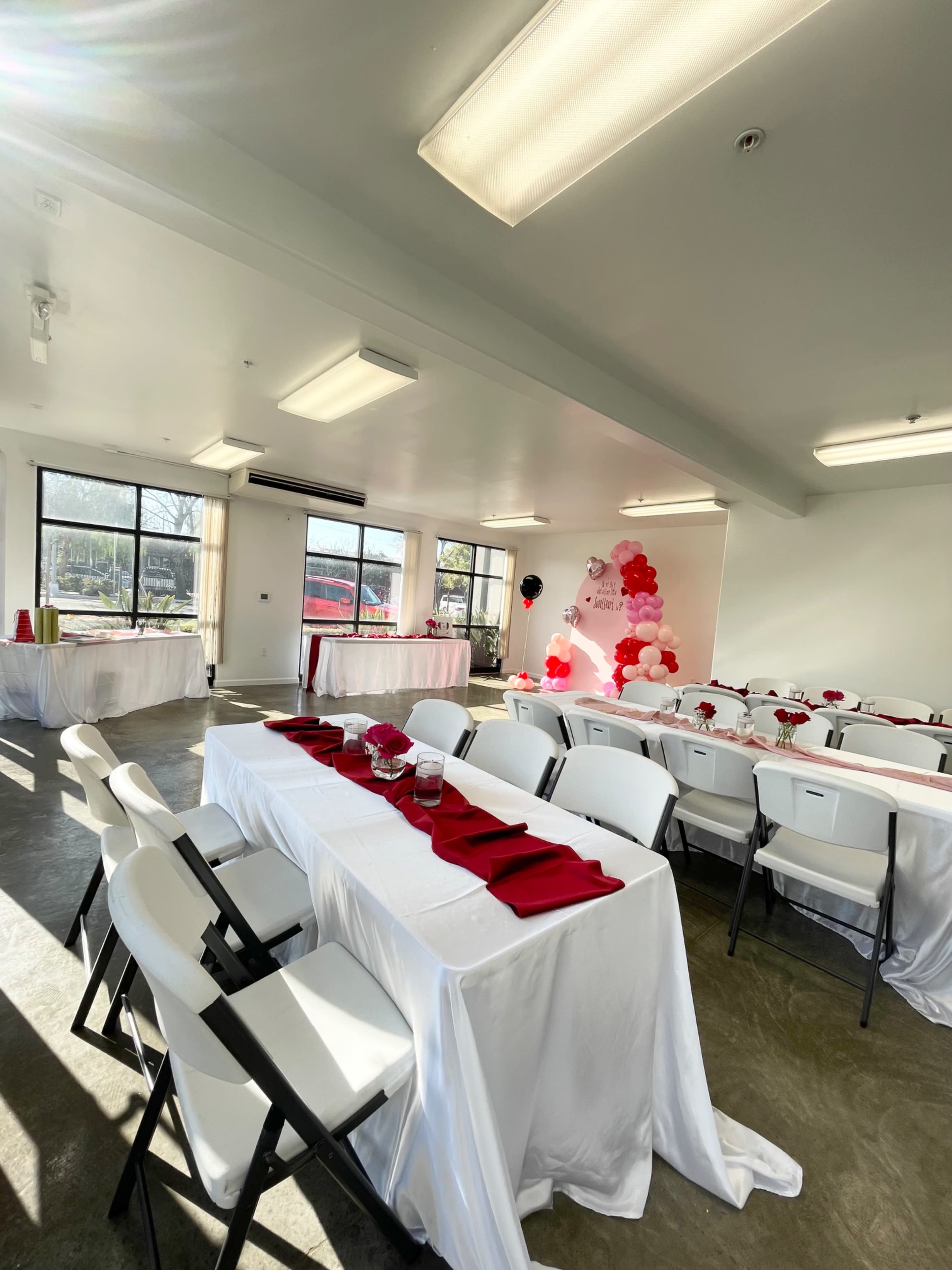 A banquet room set up with white tables covered in tablecloths, red napkins placed neatly on each setting, and a balloon decoration in the corner, all illuminated by natural light from large windows.