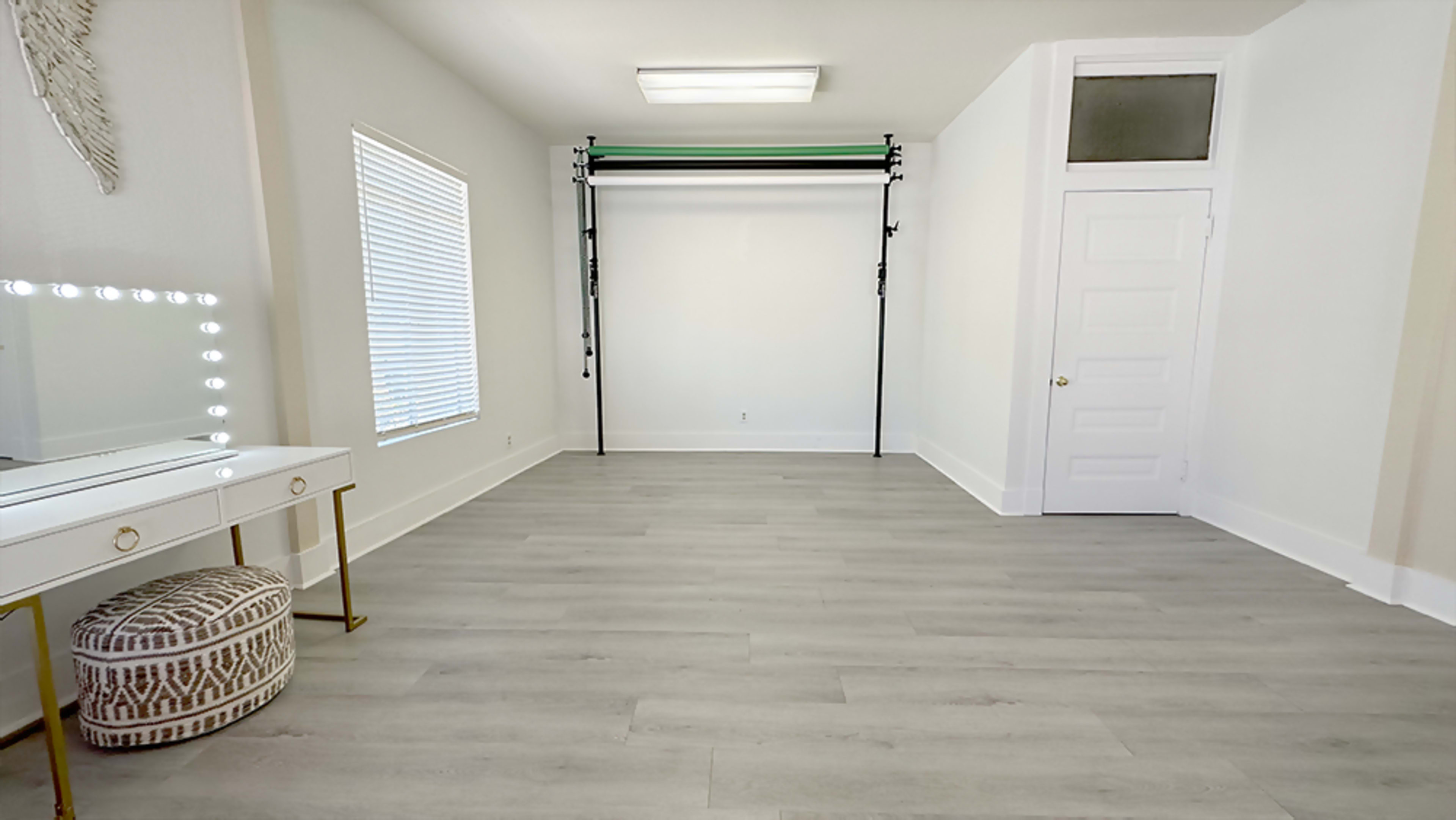 A spacious room features a large window, a folding backdrop stand, and a vanity with illuminated mirrors.