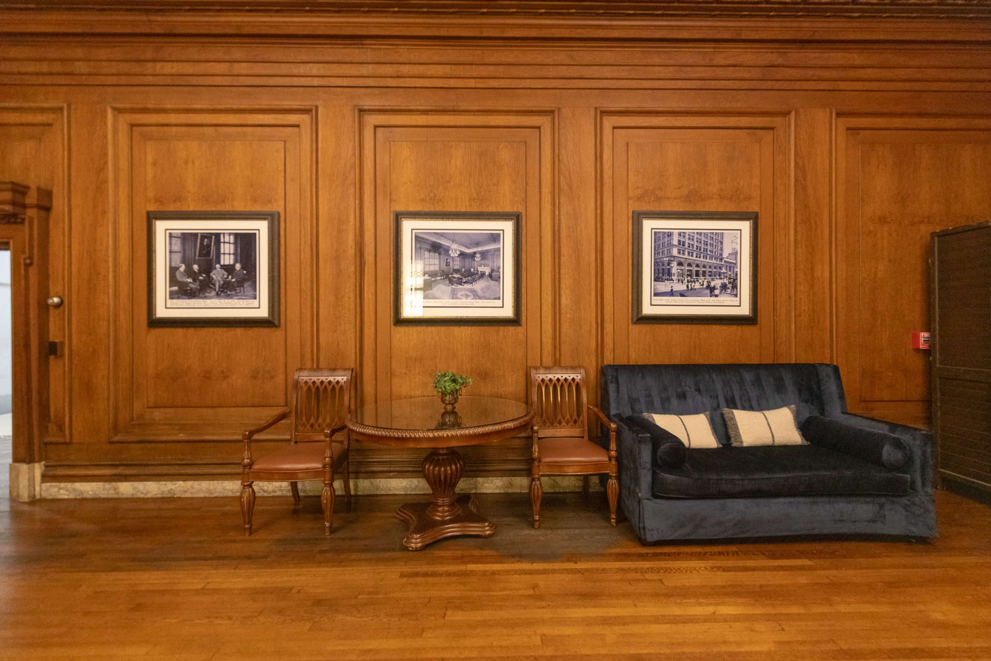 The image shows a wooden-paneled room featuring a blue sofa, two wooden chairs, and a small circular table, with framed black-and-white photographs on the walls.