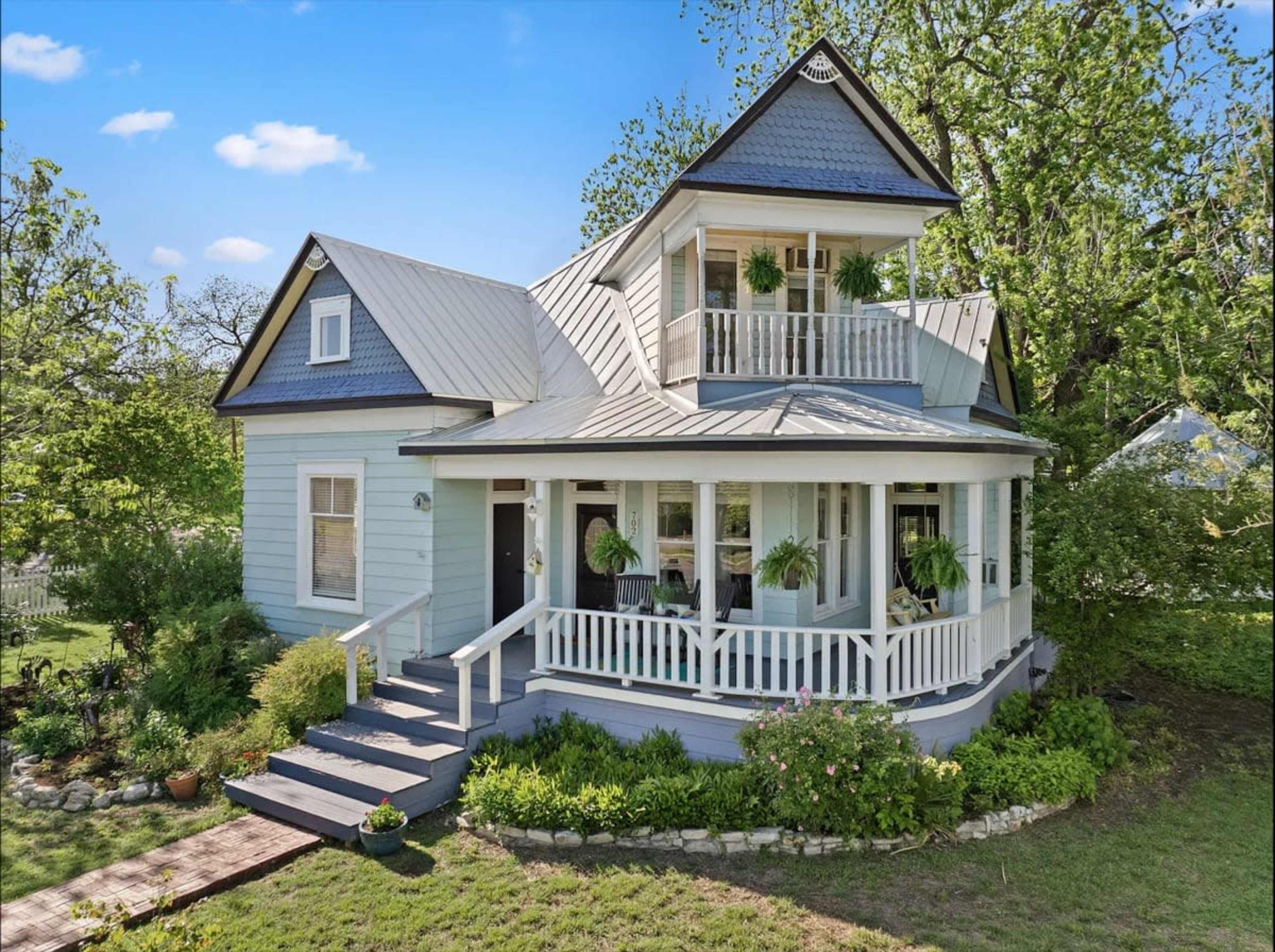 A two-story blue house with a peaked roof features a front porch adorned with potted plants and surrounded by greenery.