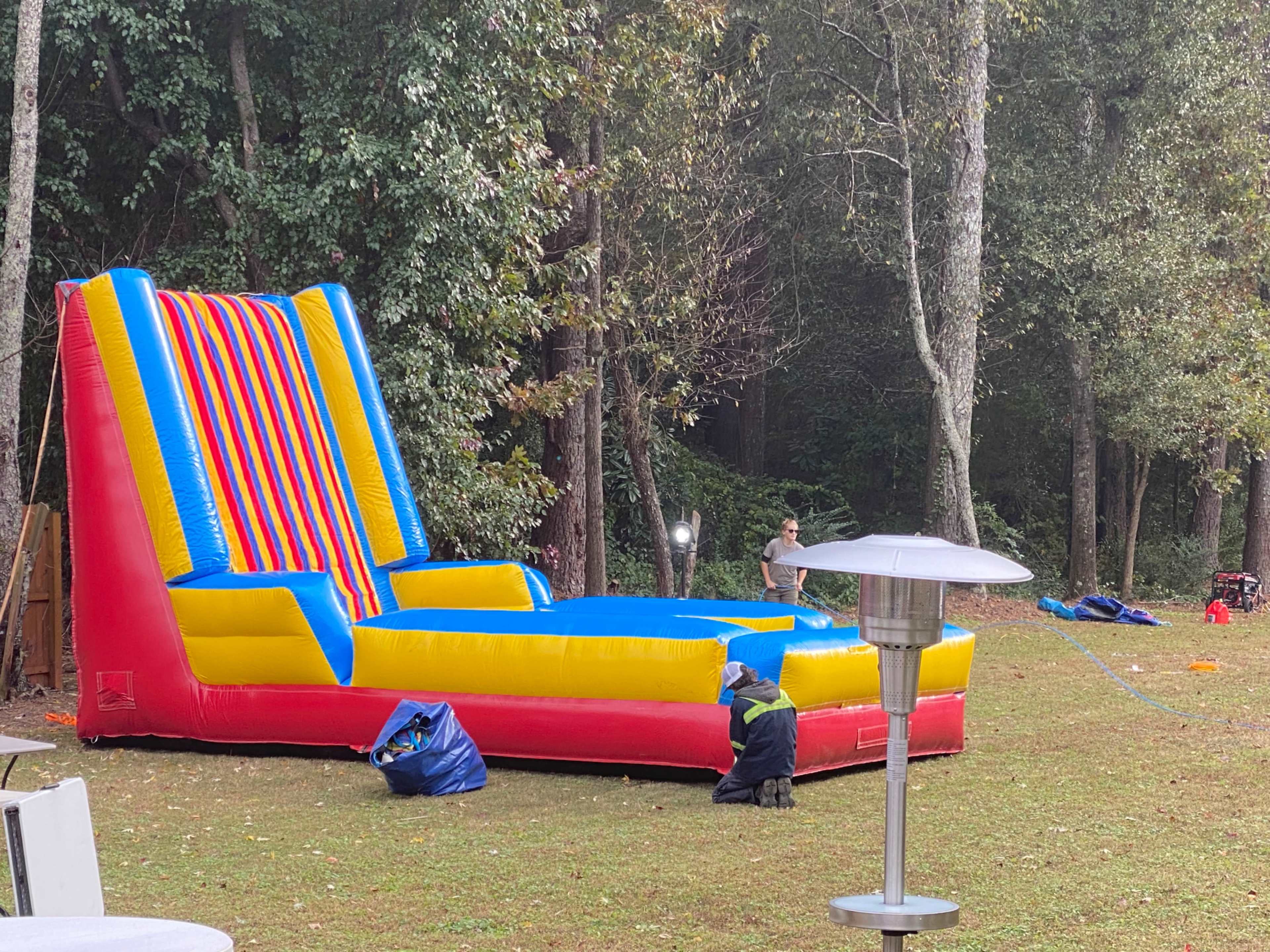 A large inflatable bounce house with colorful stripes is set up in a grassy area surrounded by trees, while a person organizes equipment nearby.