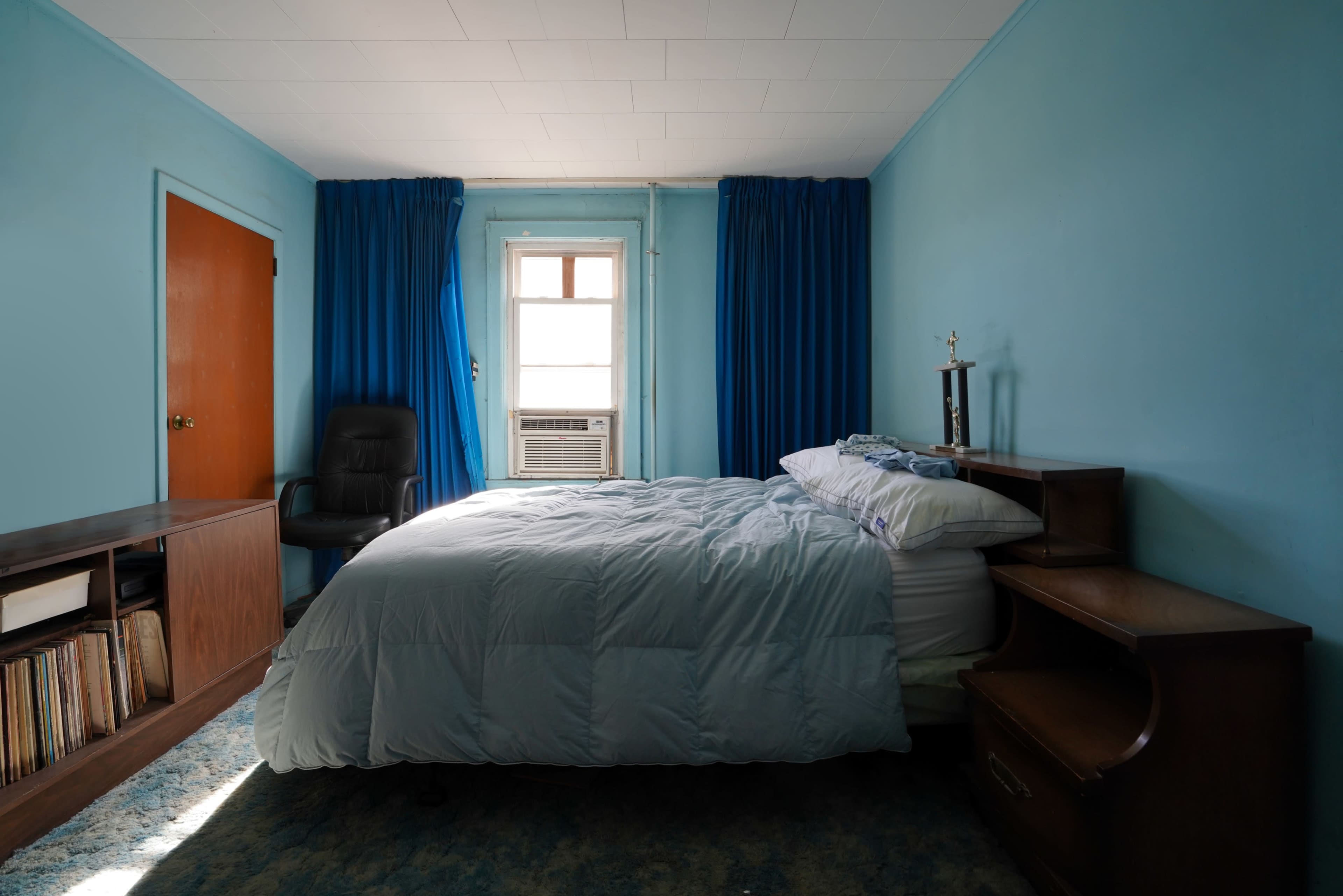 The room features a bed with a light blue comforter, a wooden nightstand, a vinyl record storage unit, a black chair, and blue curtains over the window.