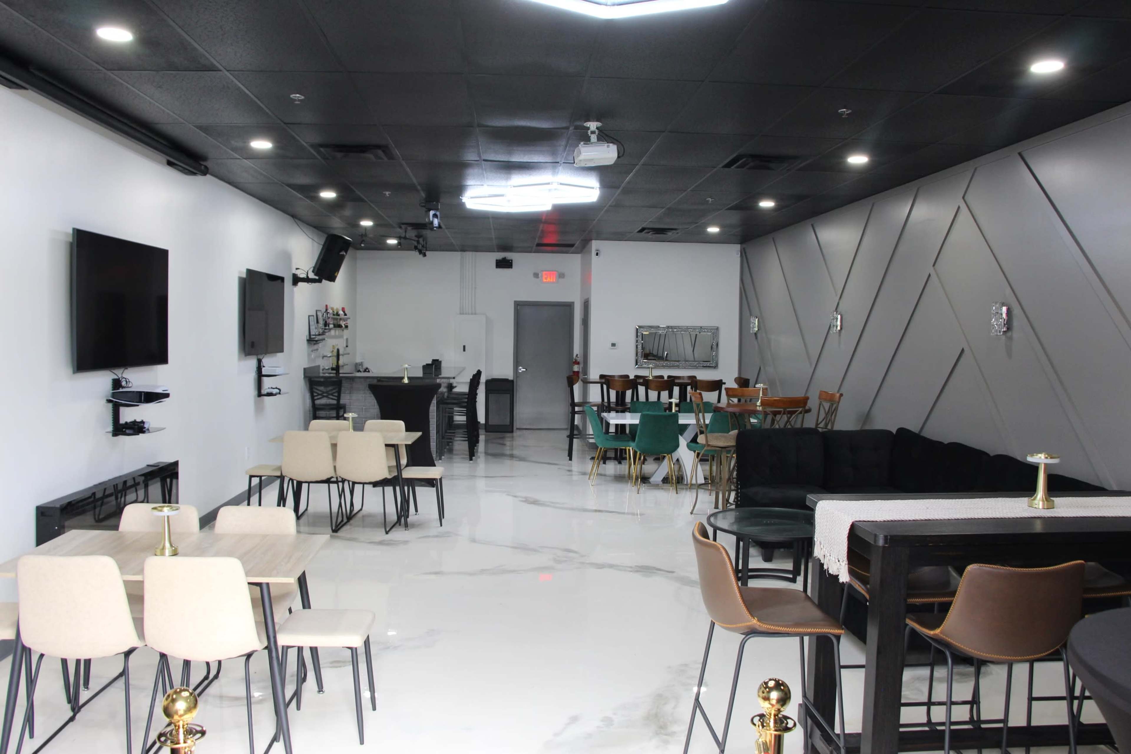 A modern lounge area featuring several tables and chairs, a bar setup, and wall-mounted televisions in a well-lit space with a polished floor.