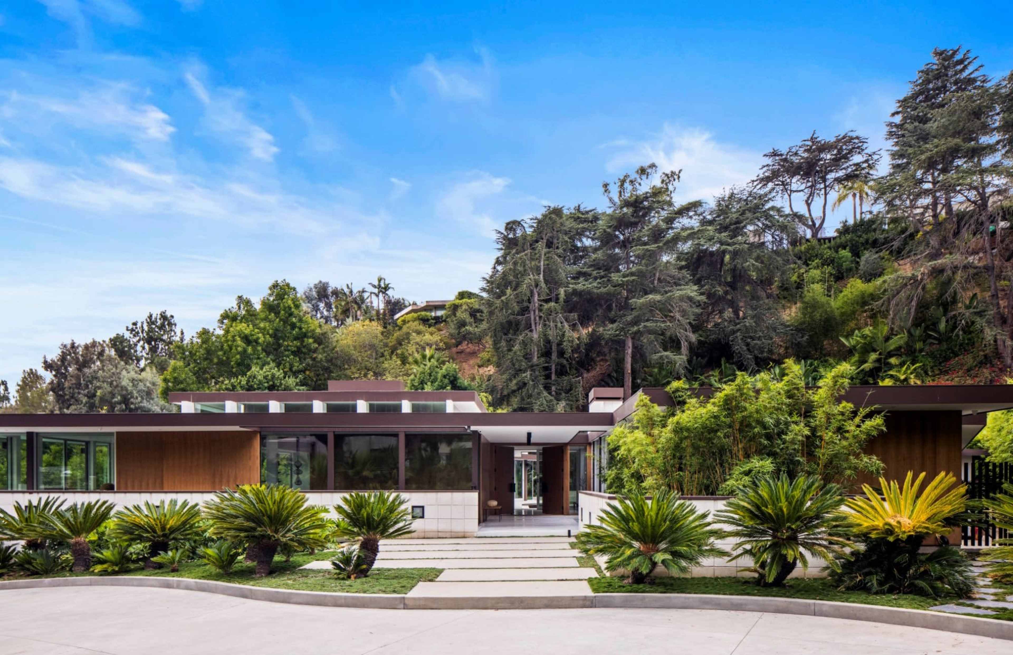 A modern house set against a hillside, surrounded by lush greenery and featuring a landscaped pathway leading to the entrance.