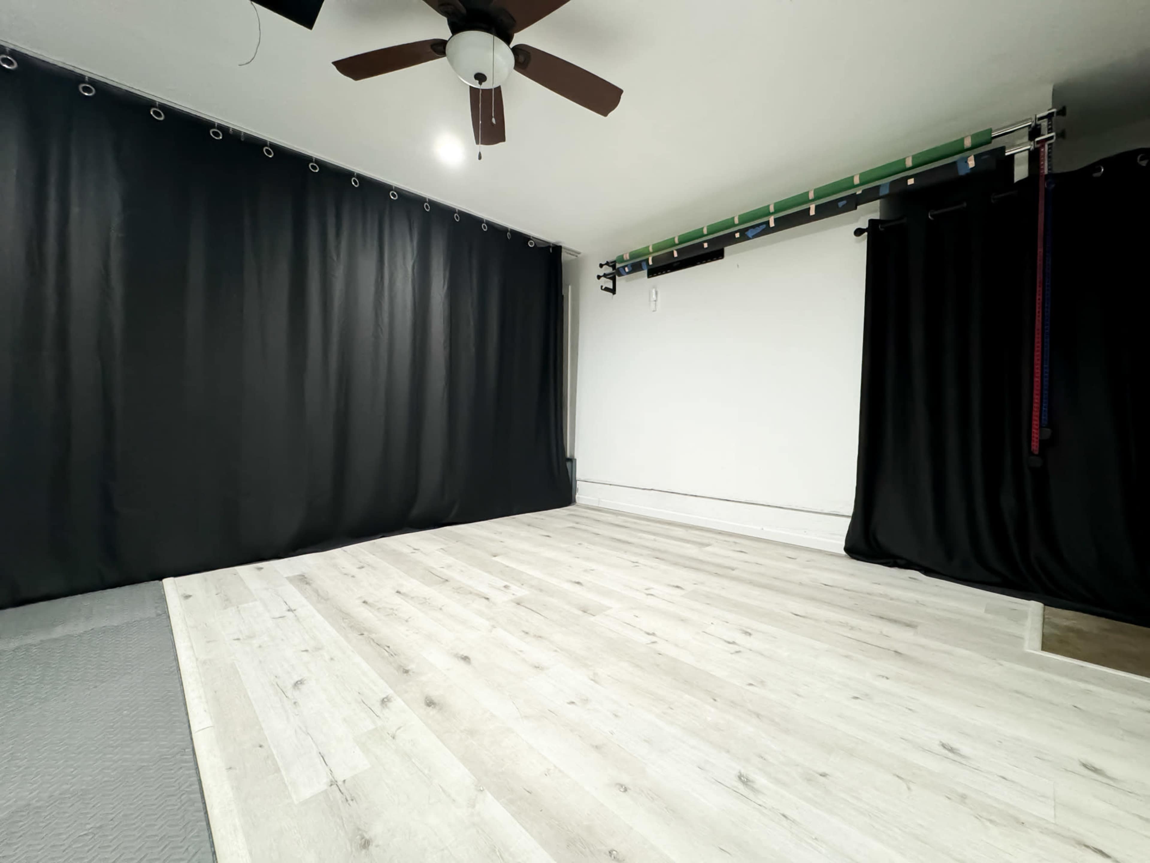 The image shows a spacious room with light wooden flooring and dark curtains covering the walls.