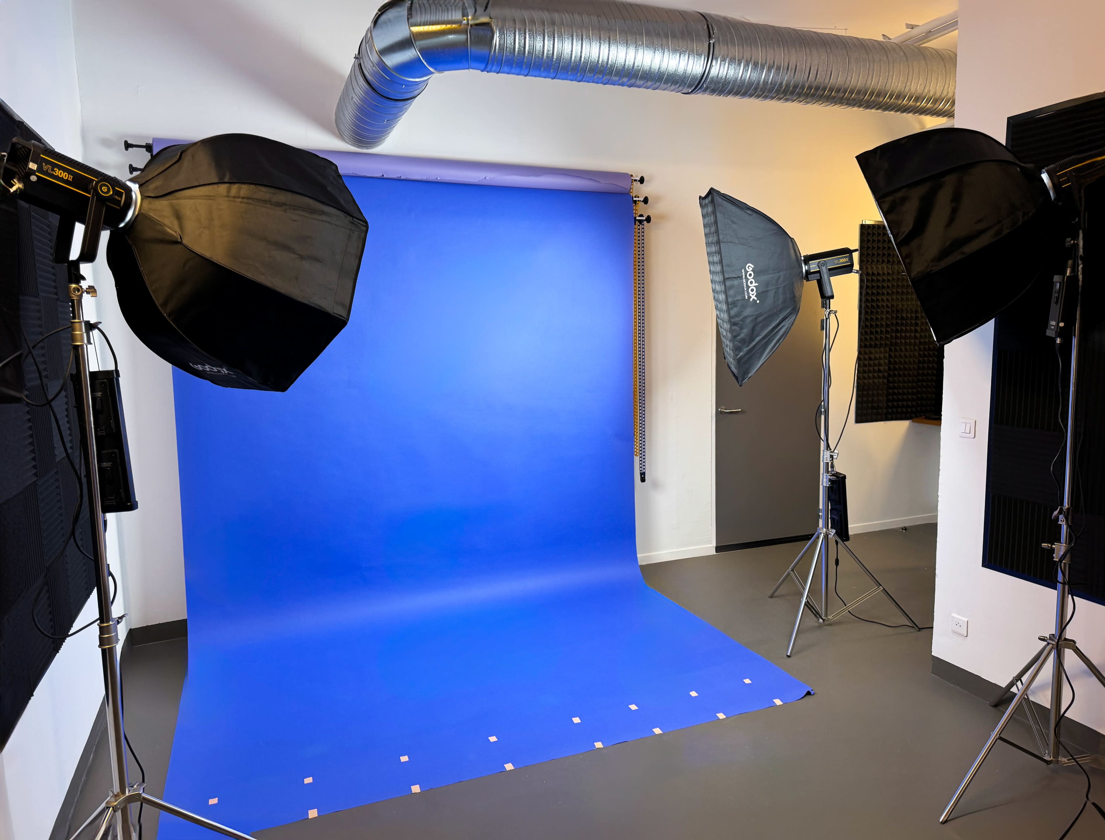 The image shows a photography studio with a blue backdrop, two softbox lights, and ventilation ducts visible on the ceiling.