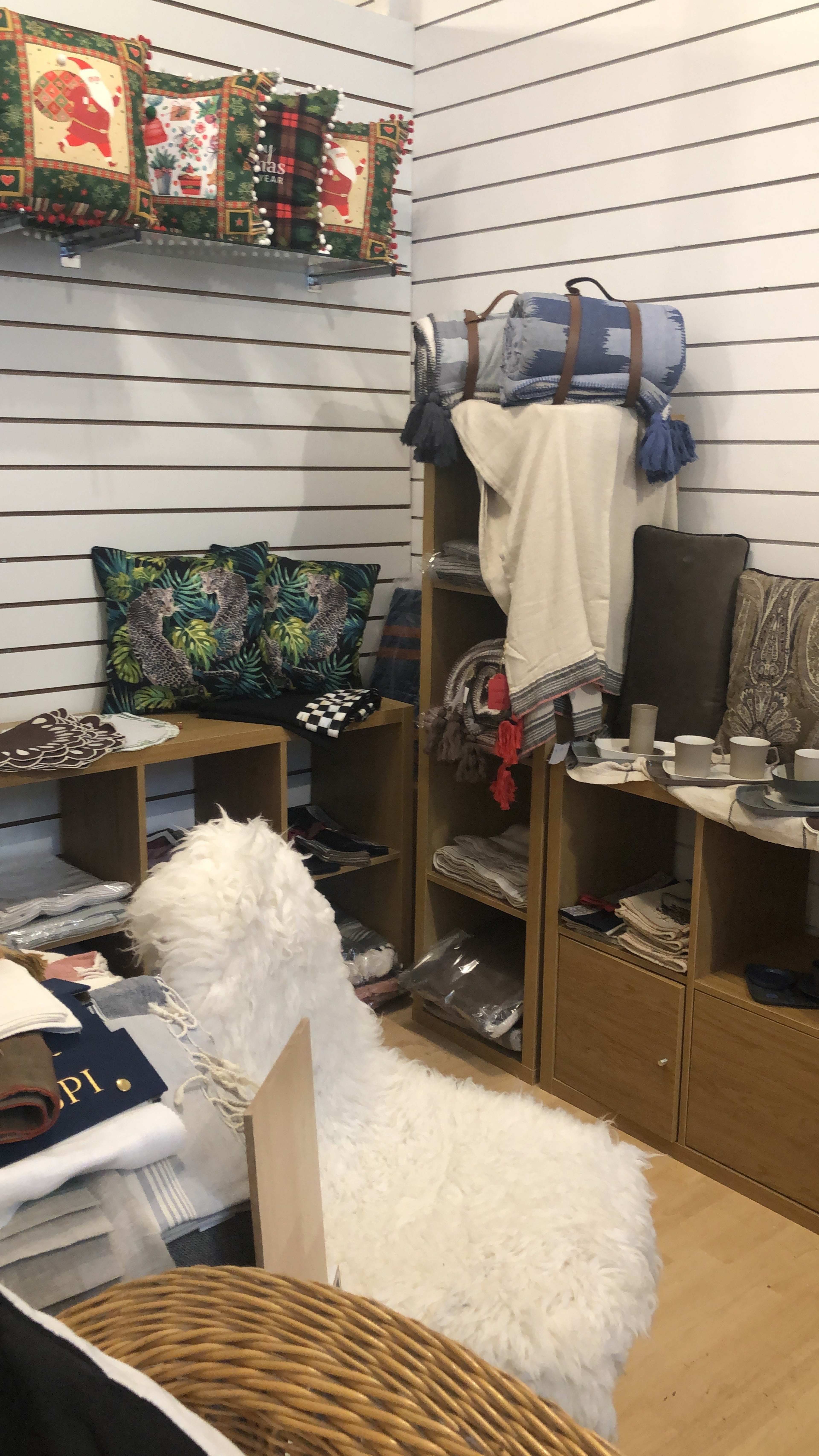 The image shows a cozy retail display featuring various home decor items, including blankets, pillows, and tableware, arranged on wooden shelves in a well-organized store corner.