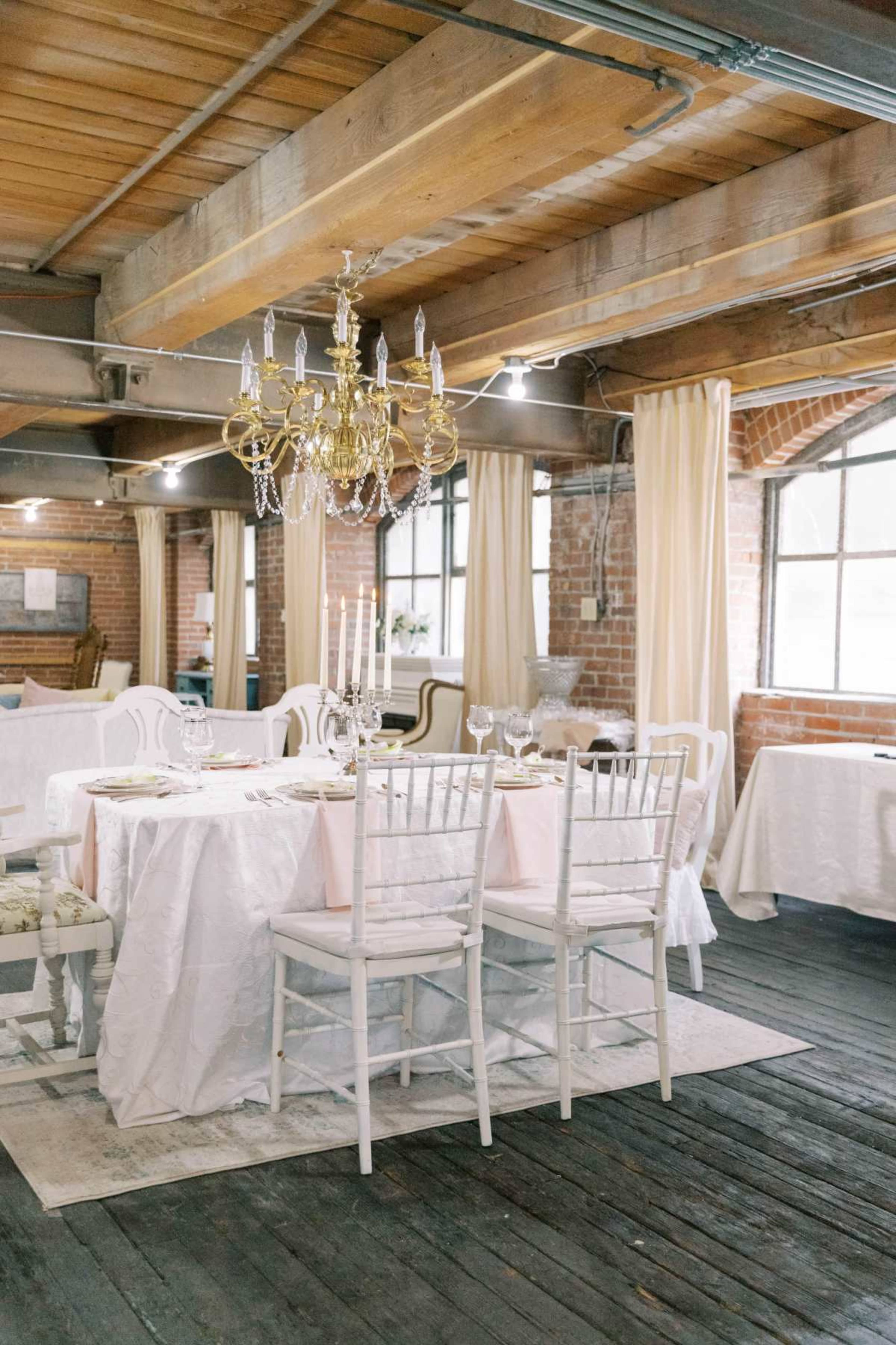 The image shows a decorated dining area featuring white tables with tablecloths, chairs, and a chandelier, set in a space with exposed brick walls and wooden beams.