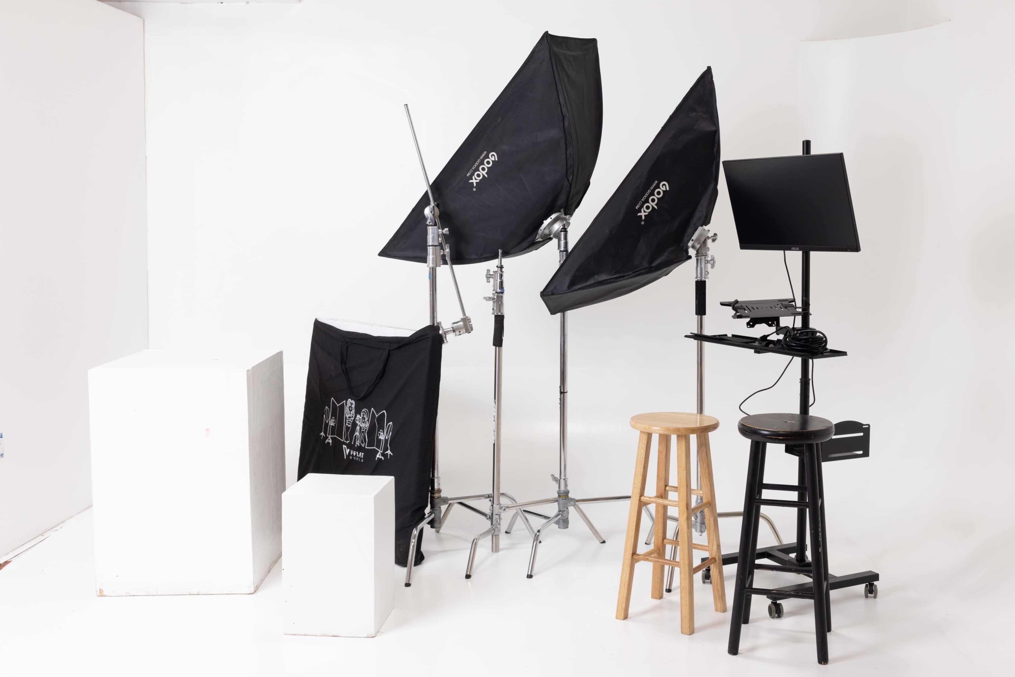 A photography setup with two softbox lights, a monitor on a stand, two stools, and two white display cubes against a white background.
