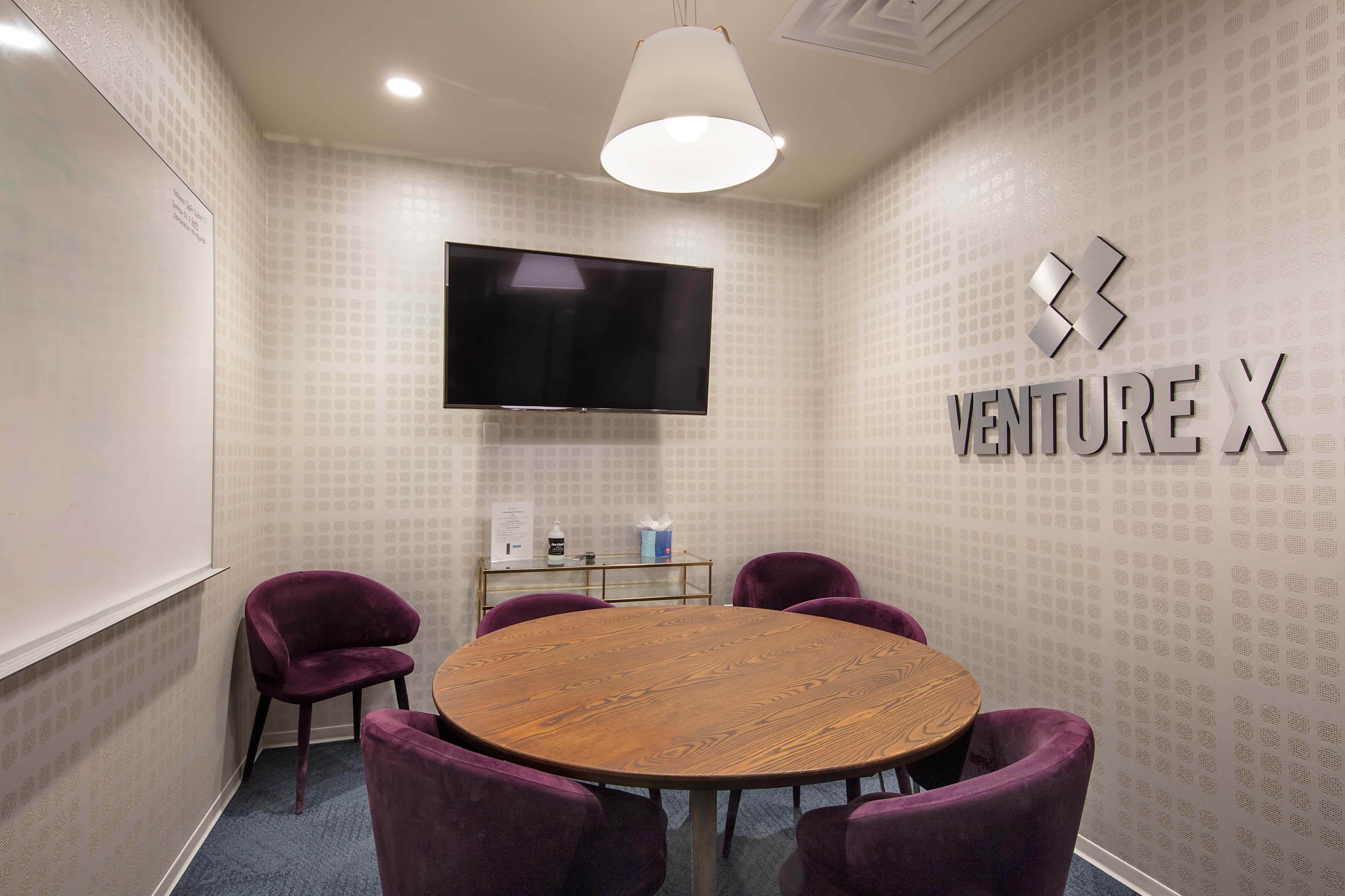 The image shows a small conference room with a round wooden table, purple chairs, a wall-mounted television, and the "VENTURE X" logo displayed on the wall.