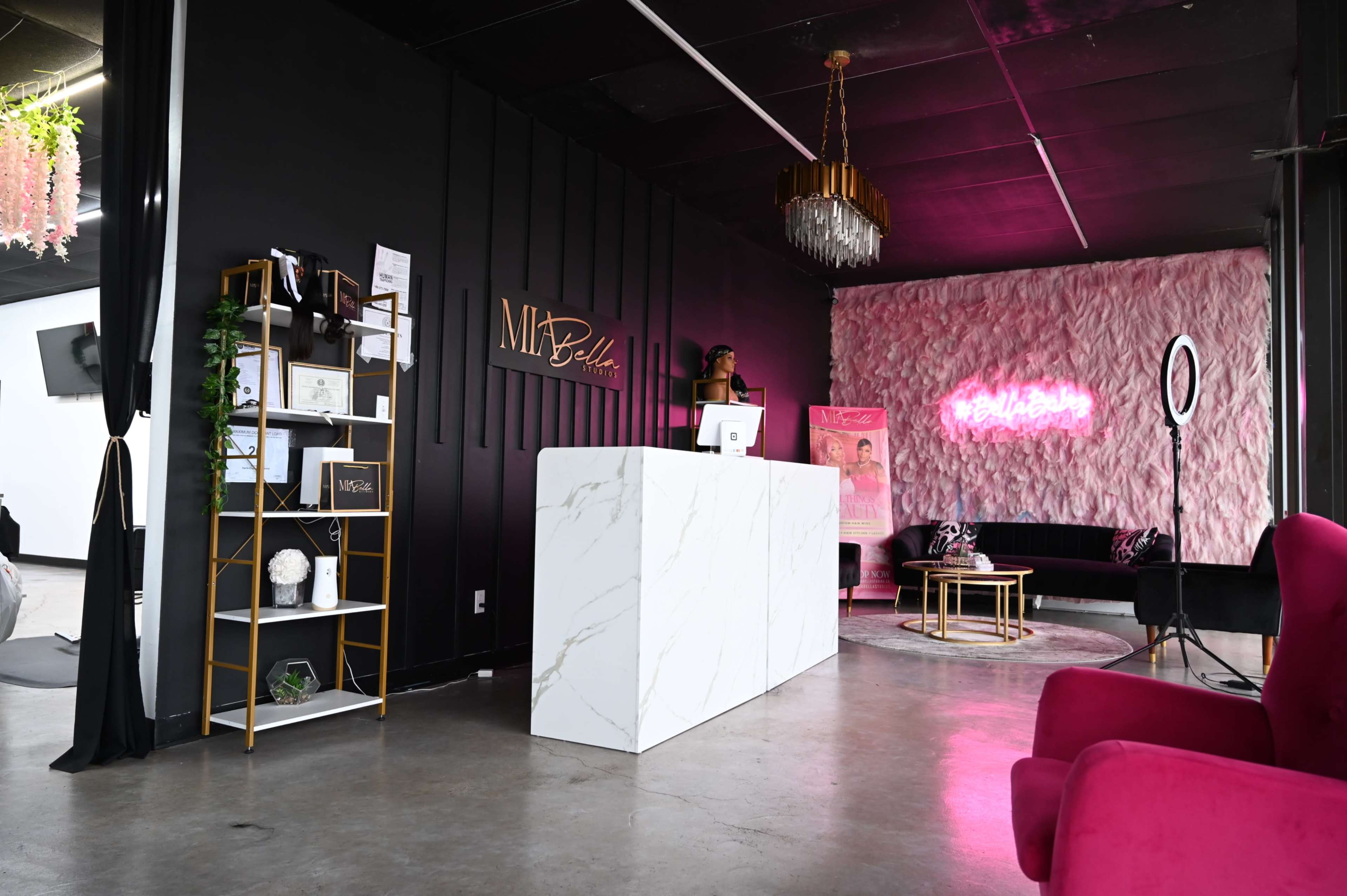 A modern reception area features a marble front desk, decorative shelving, plush seating, and a pink accent wall with neon signage.