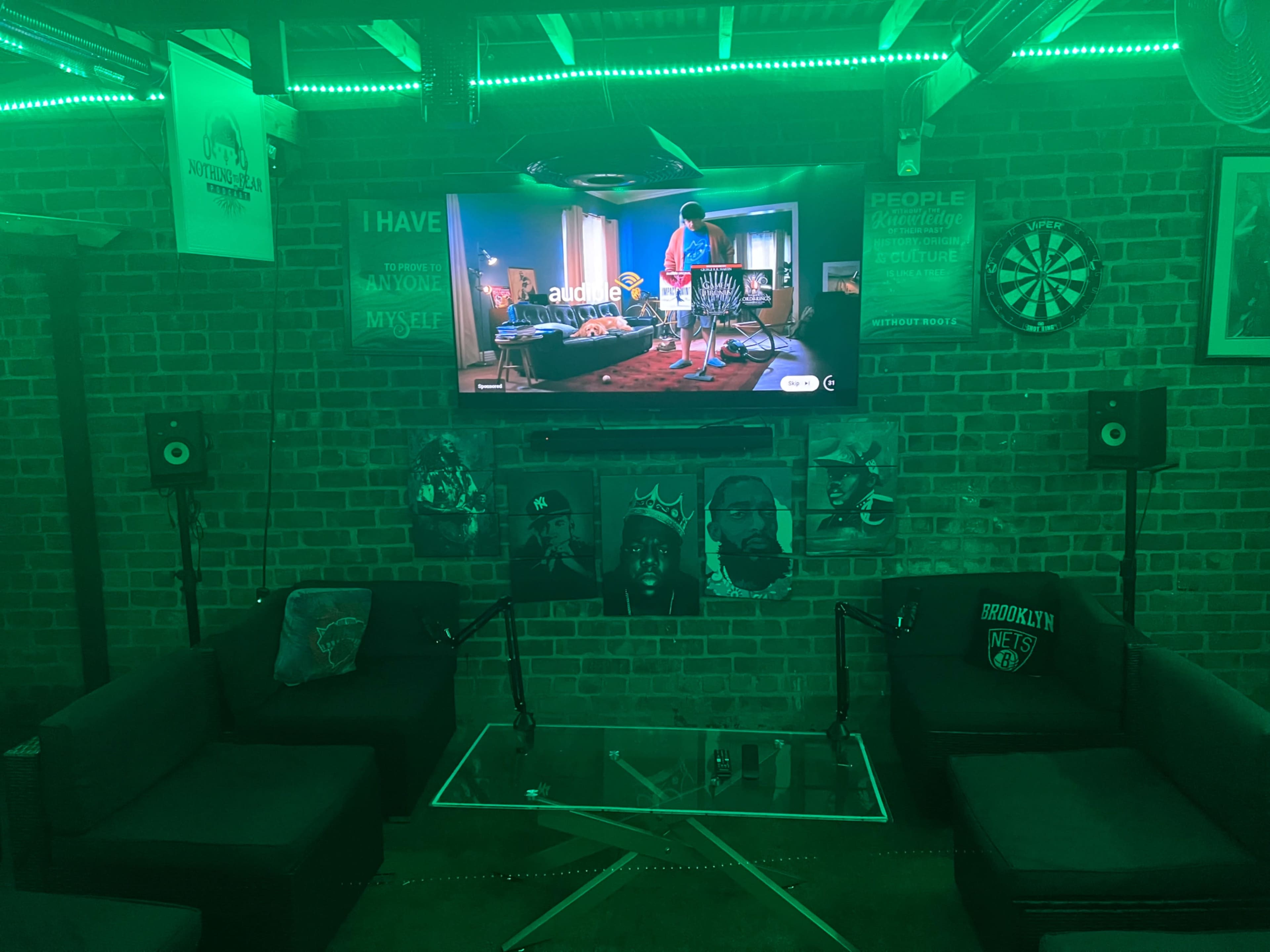 A television is mounted on a brick wall featuring artwork, with green lighting illuminating two couches and a glass coffee table in a lounge area.