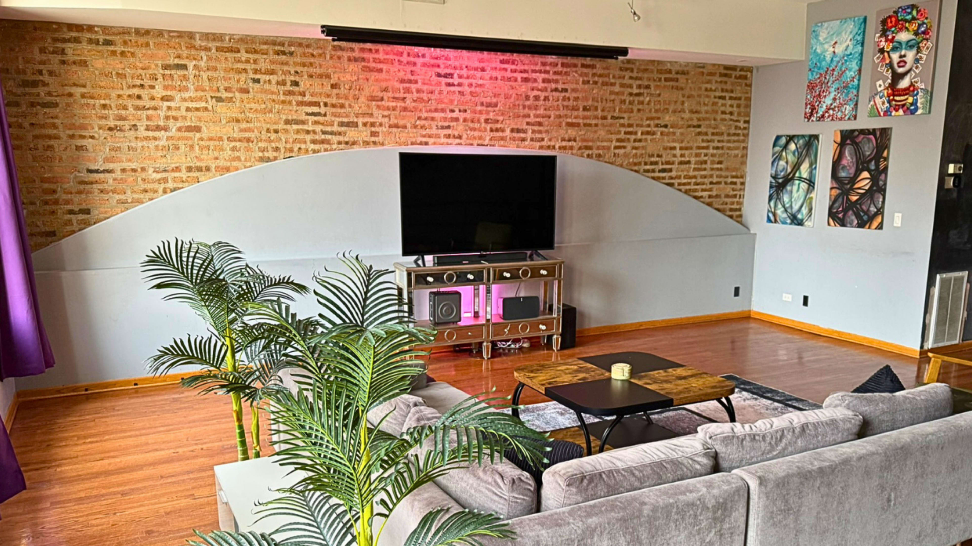 The image shows a modern living room with a large TV mounted on a brick wall, a gray sofa, a coffee table, and colorful artwork on the walls.