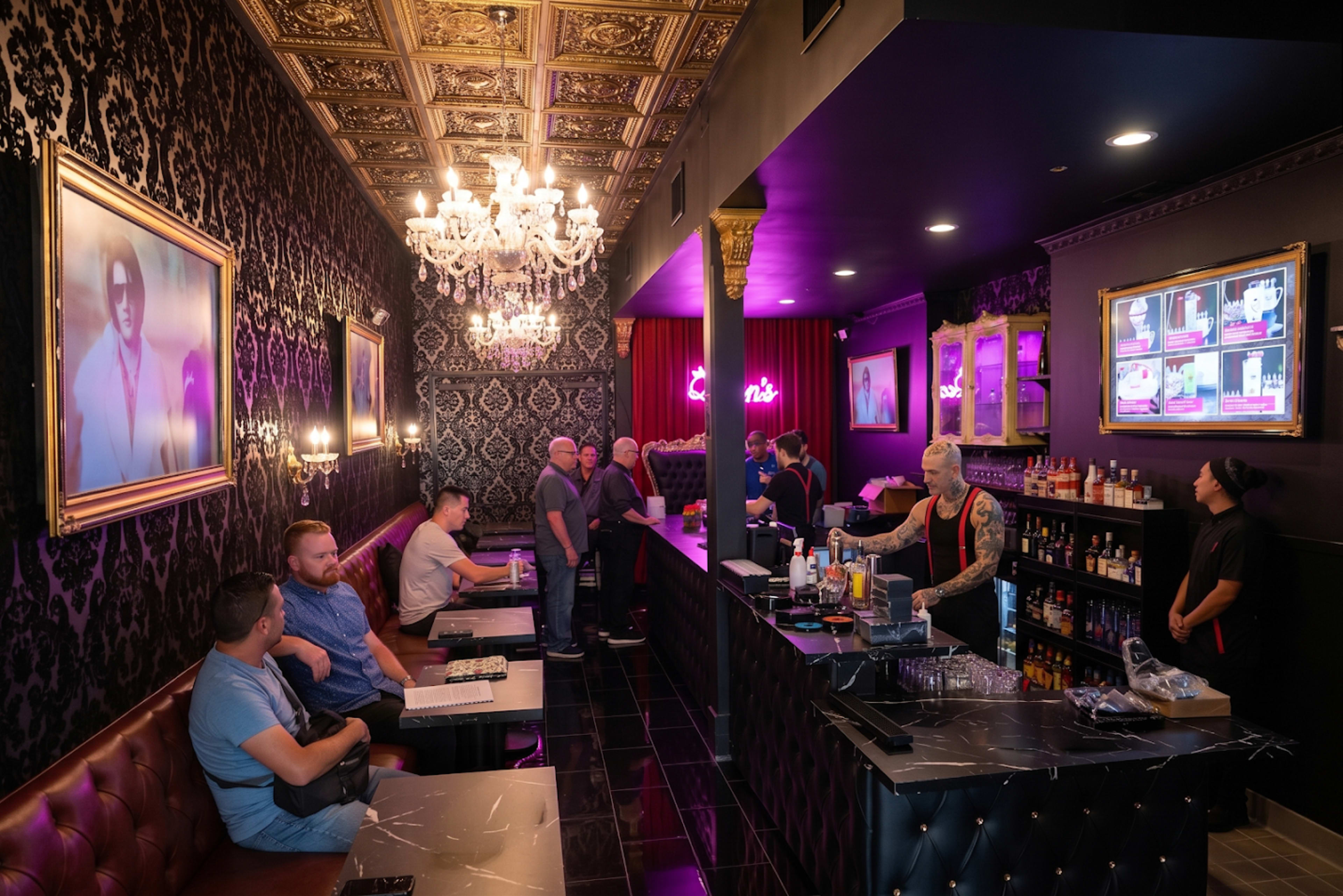 The image shows a stylish bar interior with dark decor, featuring patrons seated at tables and a bartender preparing drinks behind a sleek counter.