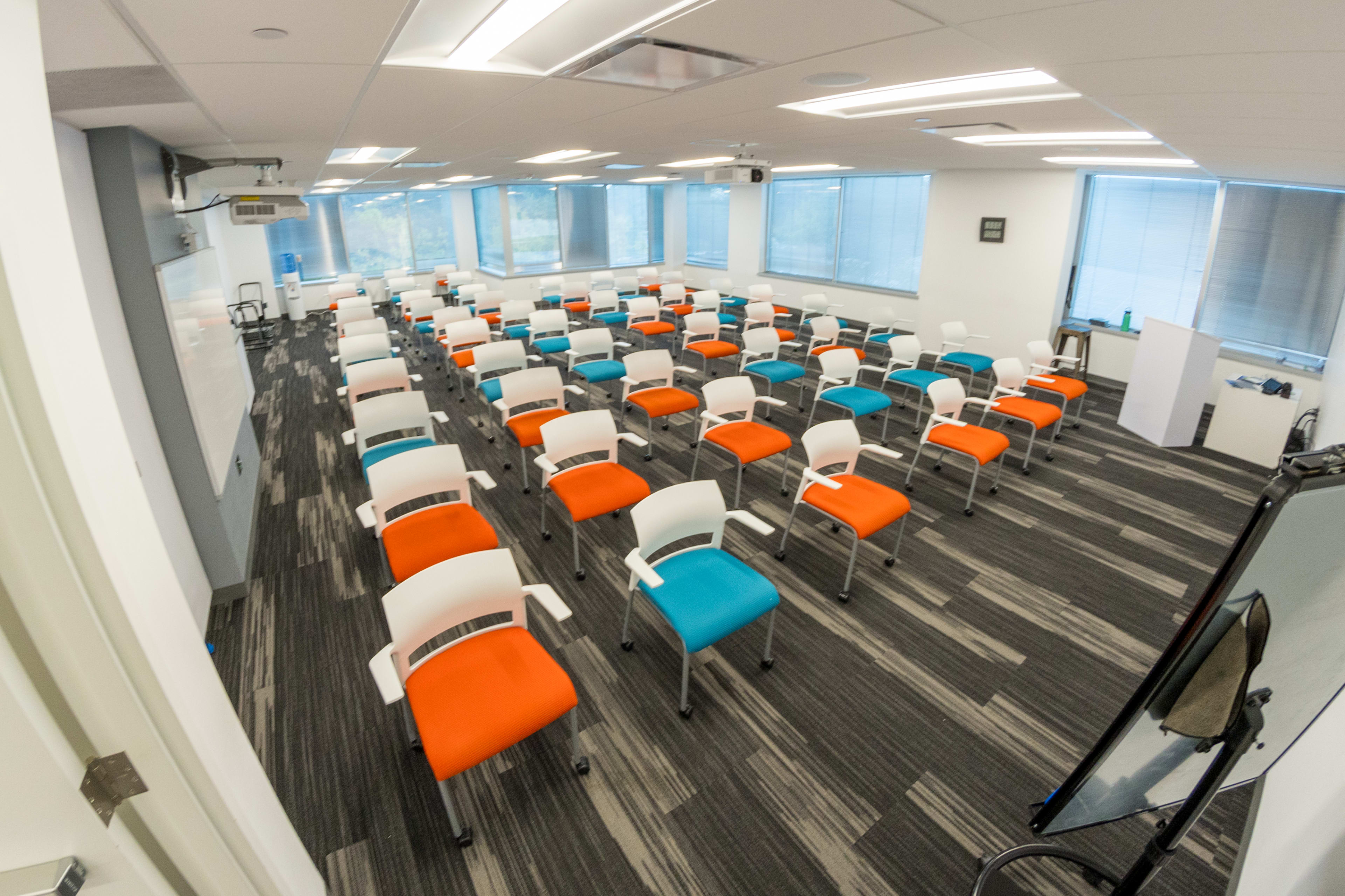 The image shows a modern classroom arranged with rows of multi-colored chairs and large windows providing natural light.