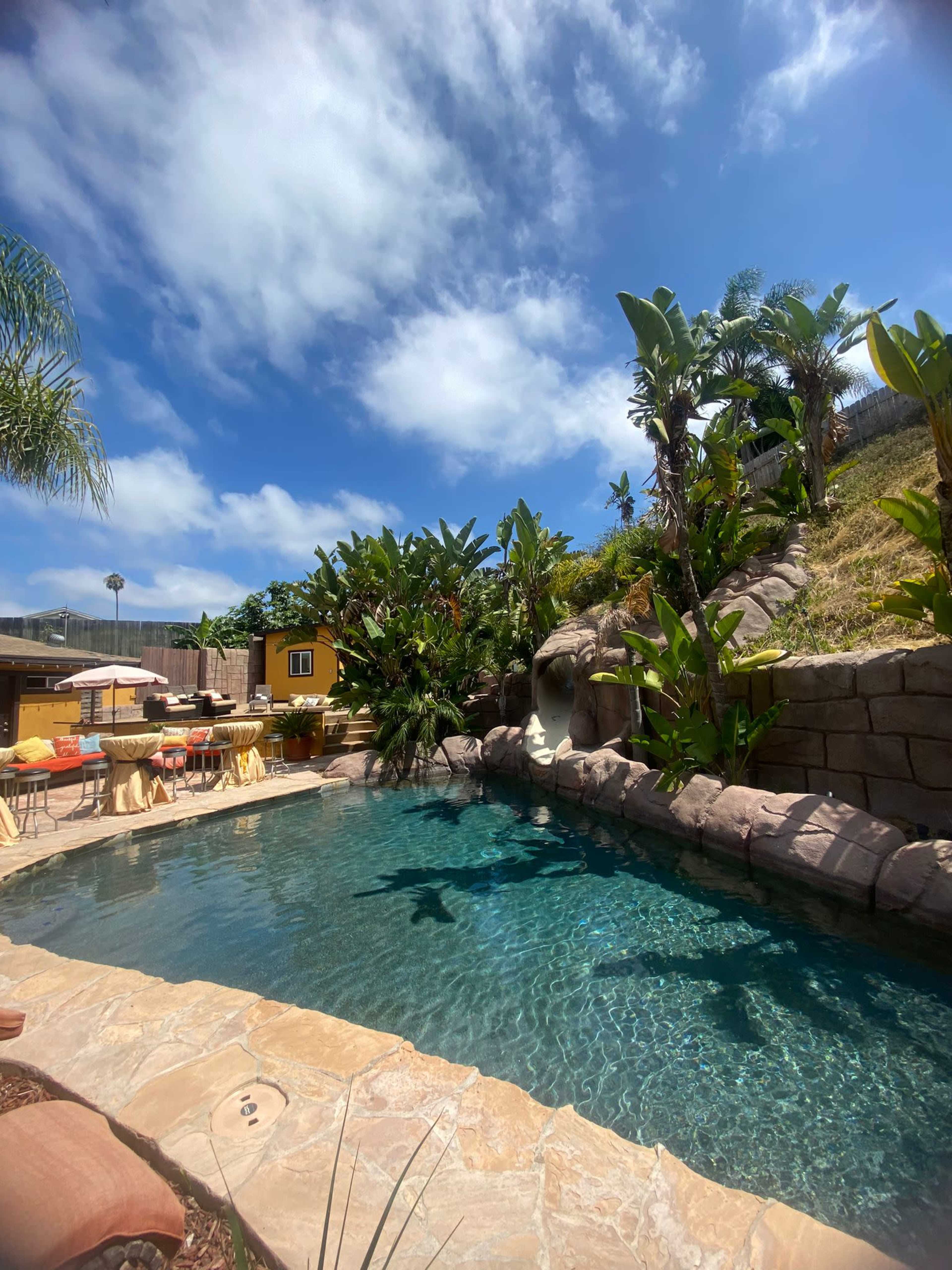 A crystal-clear pool surrounded by tropical plants and a rocky landscape under a blue sky with scattered clouds.
