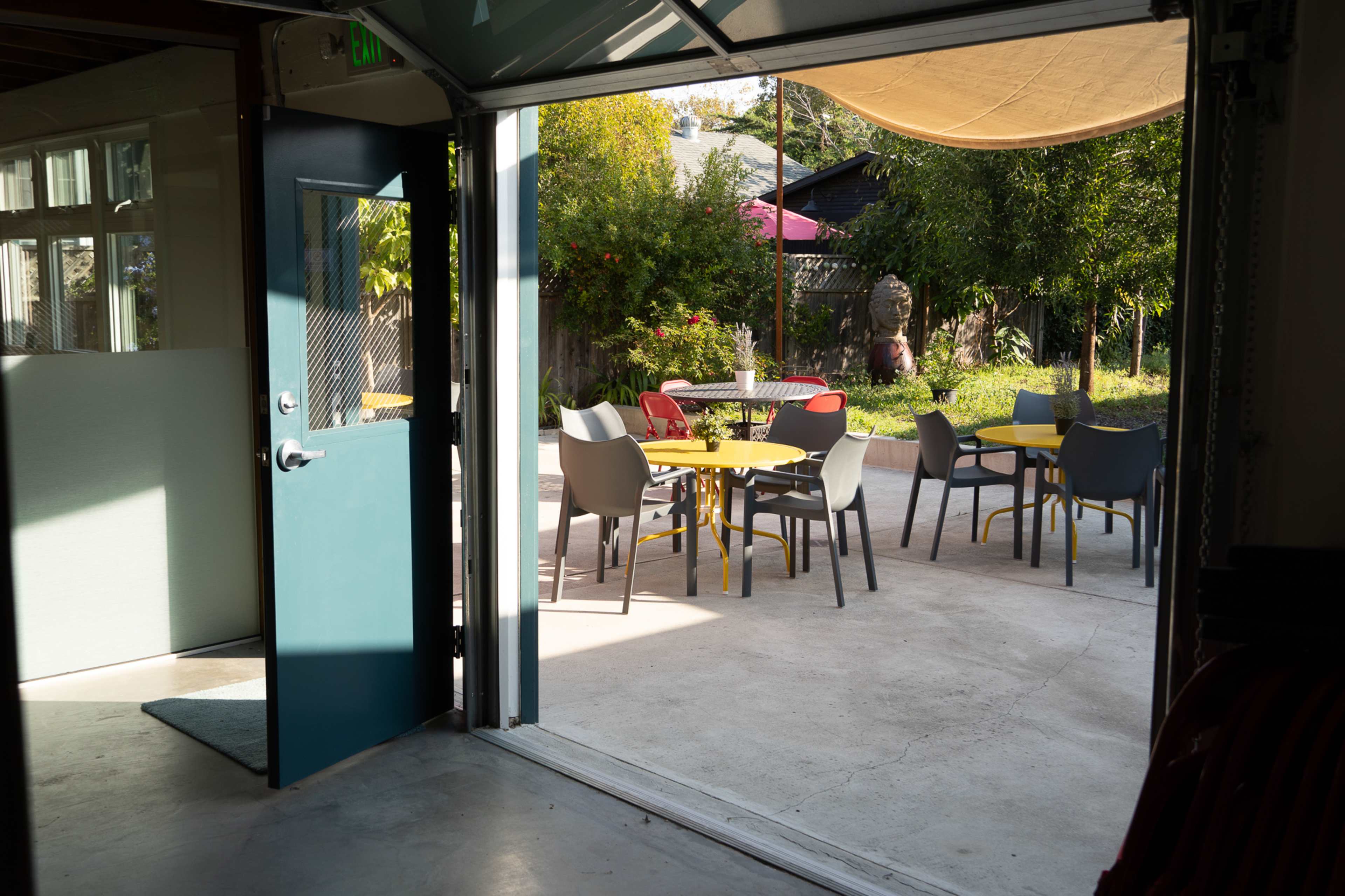 An open door leading to an outdoor seating area with yellow tables and various chairs in a garden setting.