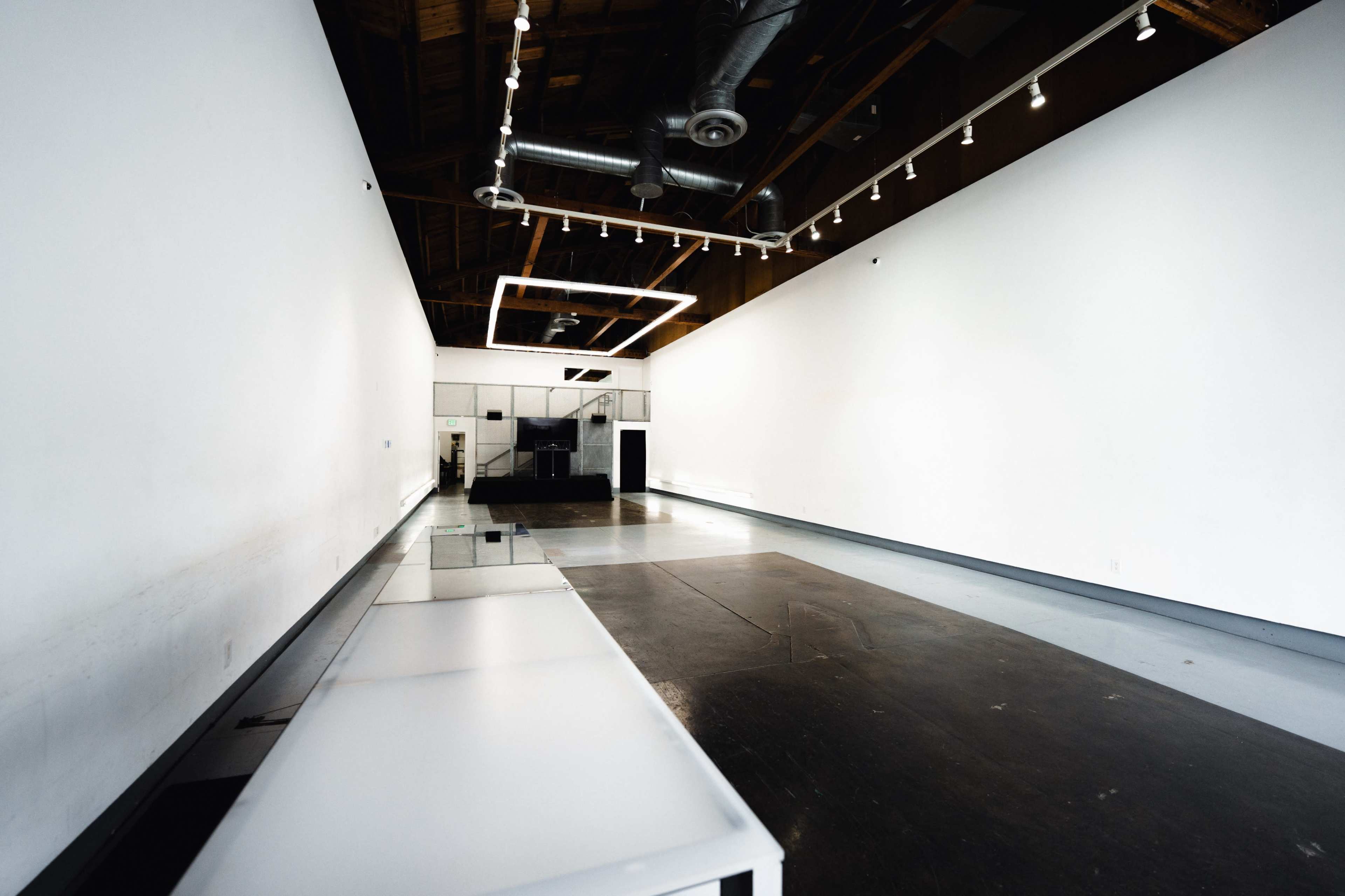 The image shows a spacious, empty interior room with white walls, a long white table on the left, and dim lighting fixtures along the ceiling.