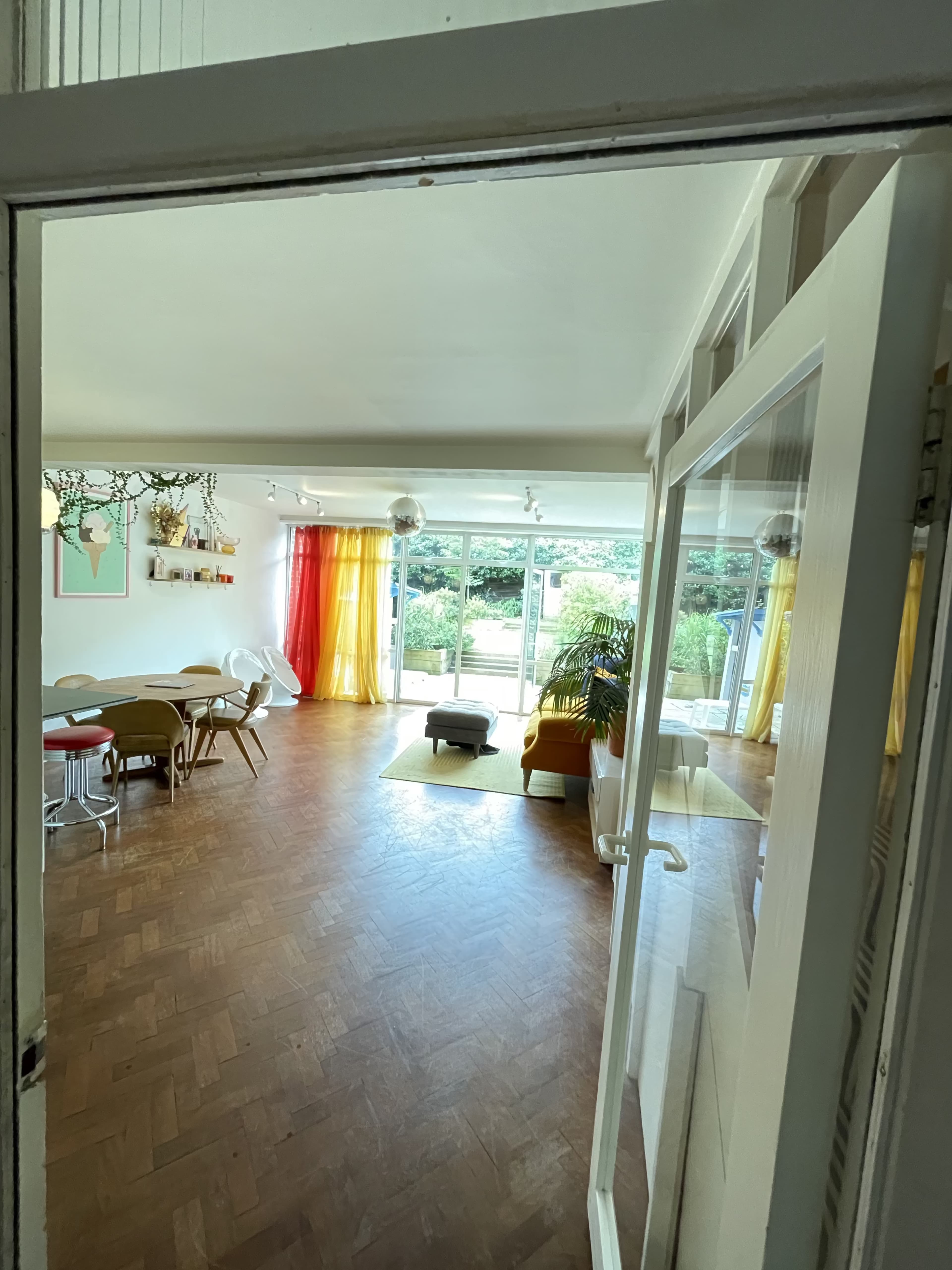 A bright living space features large windows with colorful curtains, a mix of modern furniture, and access to a garden.