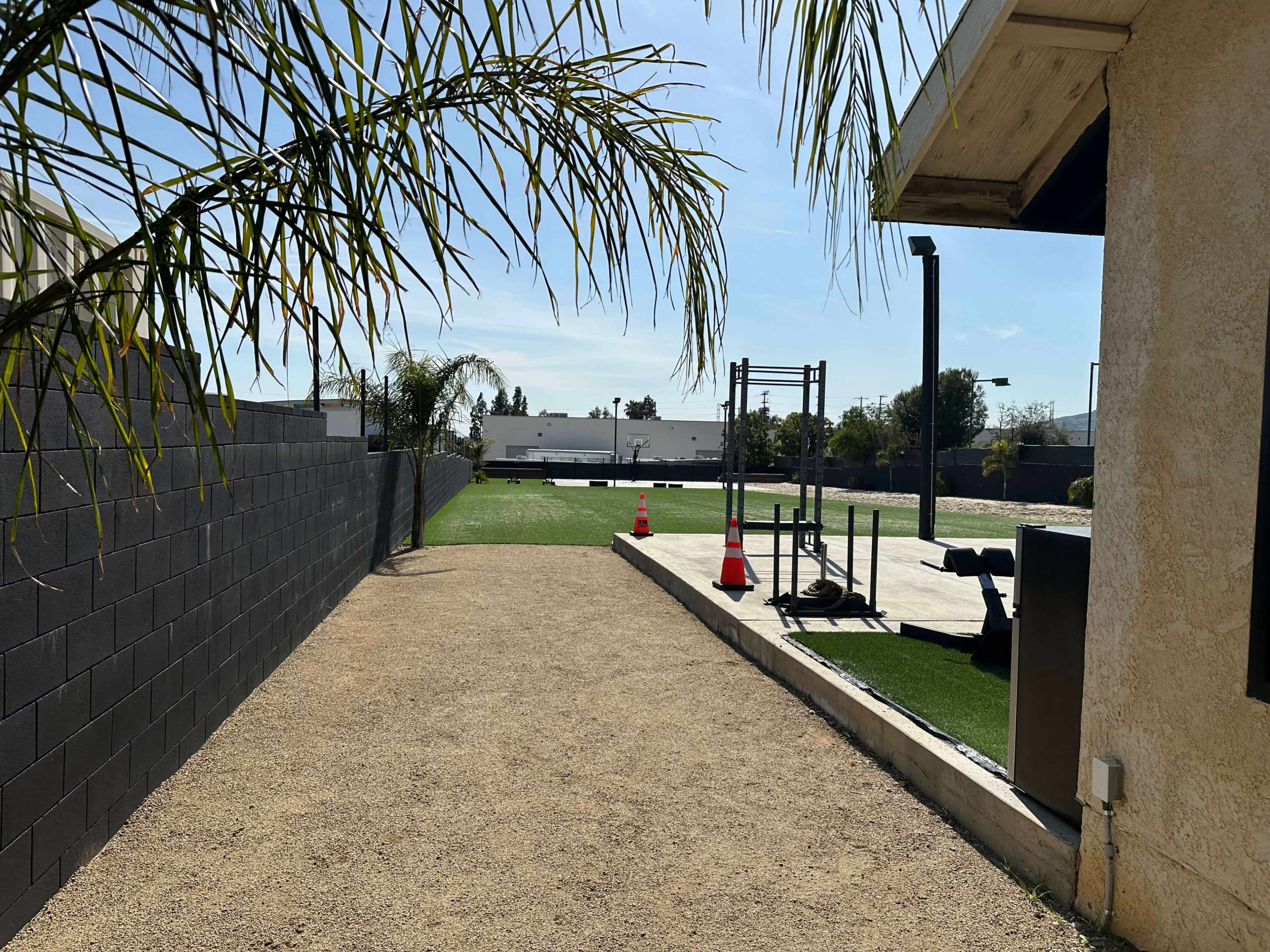 The image shows a narrow pathway alongside a wall, leading to a grassy area with exercise equipment in the distance.