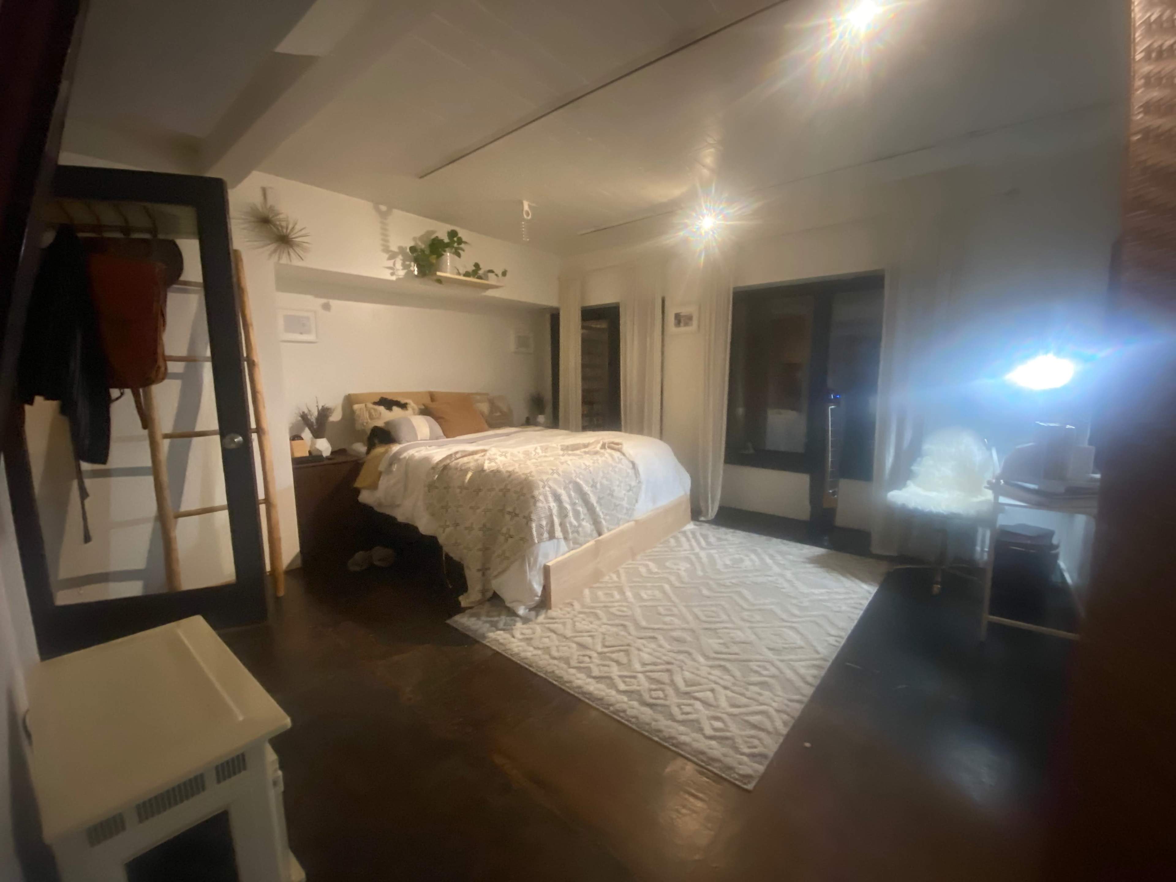 A well-furnished bedroom features a large bed with a decorative blanket, a desk with a lamp, and a plush rug on the dark wooden floor.