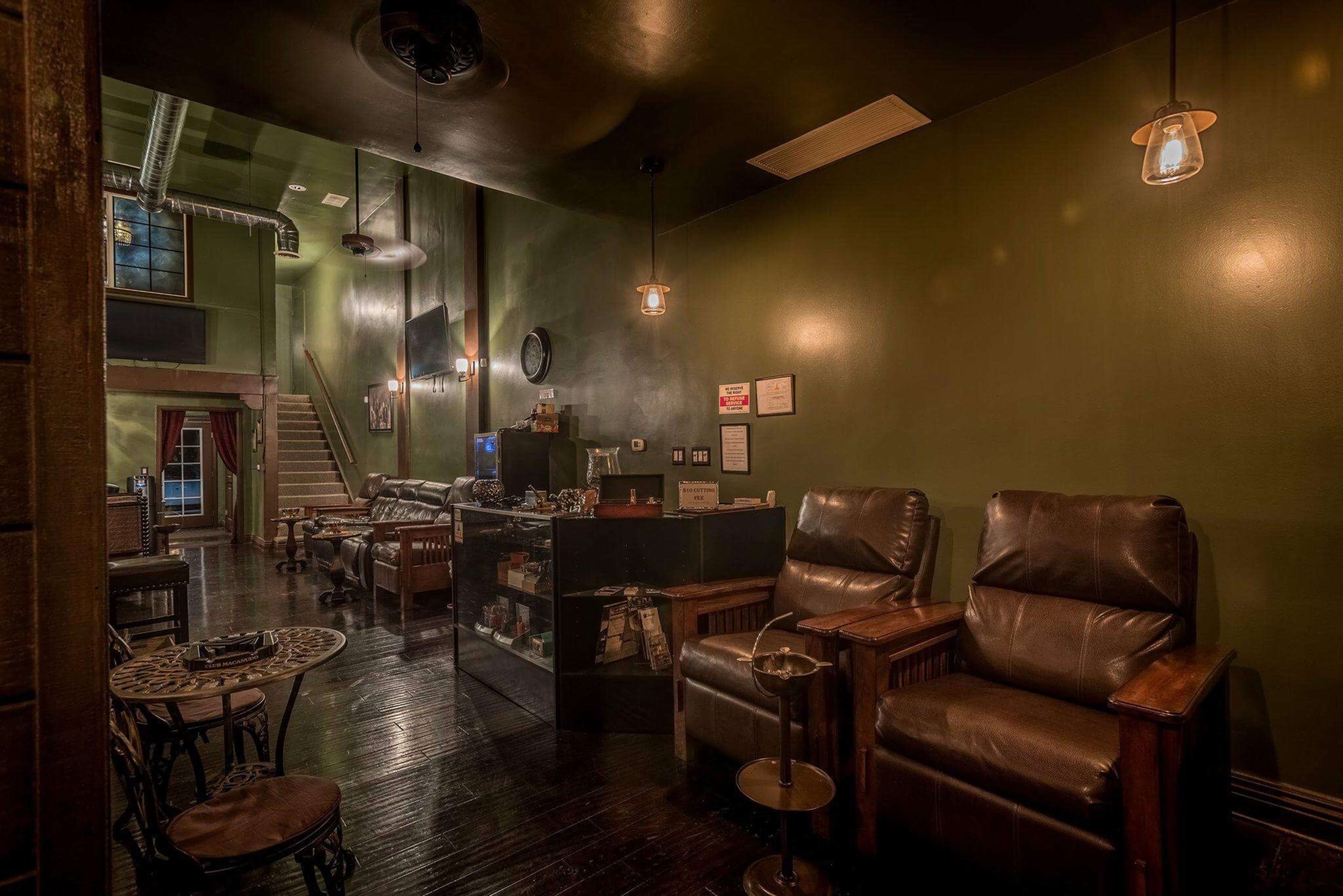 A dimly lit lounge with dark green walls, leather armchairs, and a staircase at the back, featuring a display counter and various furnishings.