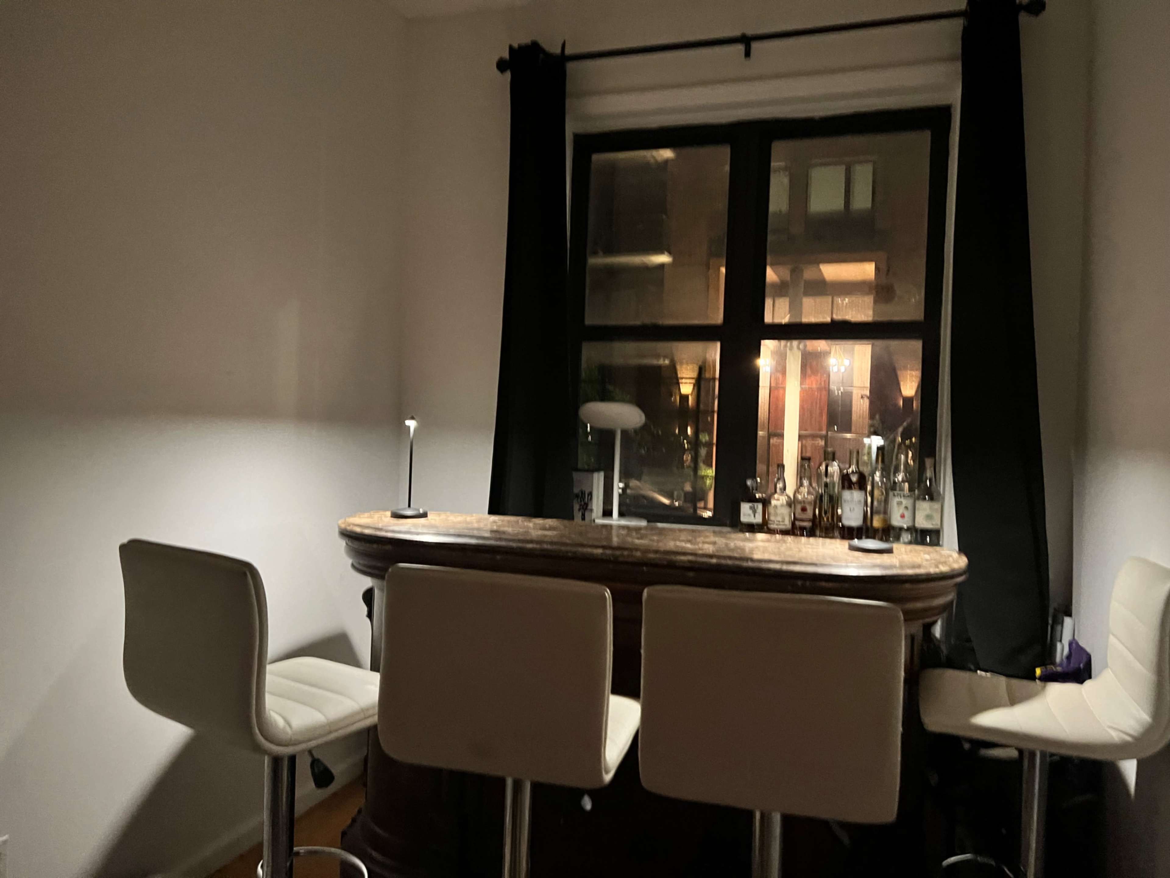 A well-lit home bar with a wooden counter and several bottles displayed on the shelf, accompanied by four white bar stools.