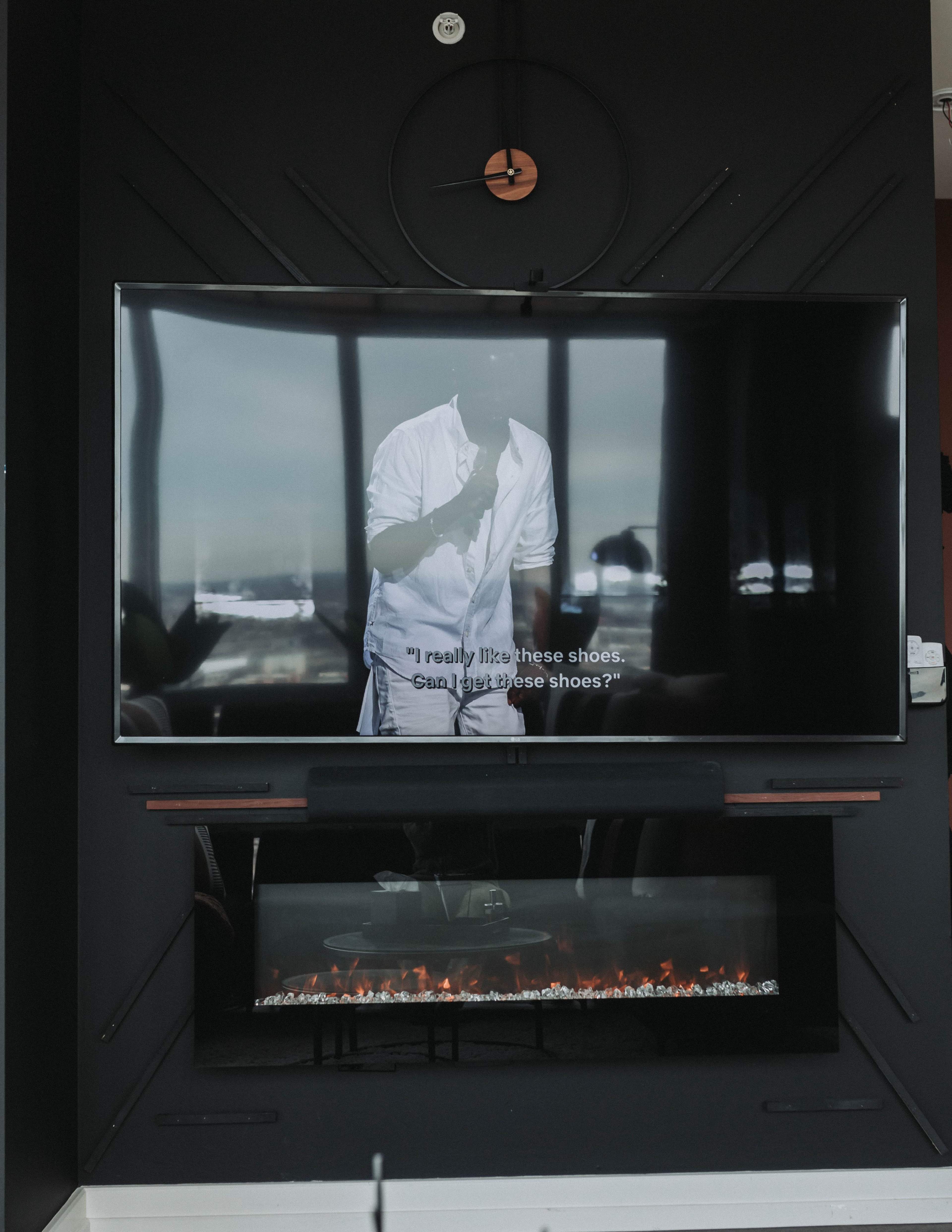 A large television displaying a person speaking, with subtitles, is mounted on a black wall above a modern electric fireplace.