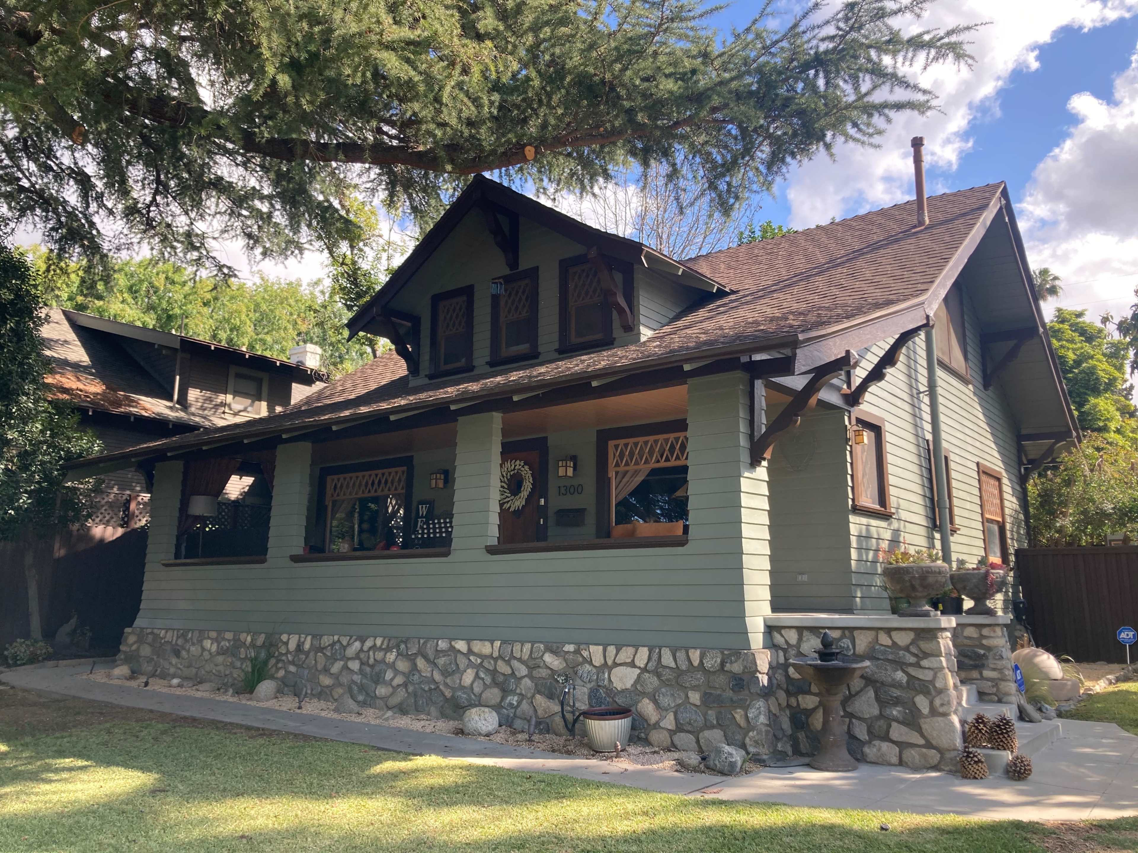 The image shows a two-story, green wooden house with a stone foundation and a front porch, surrounded by grassy landscaping and trees.