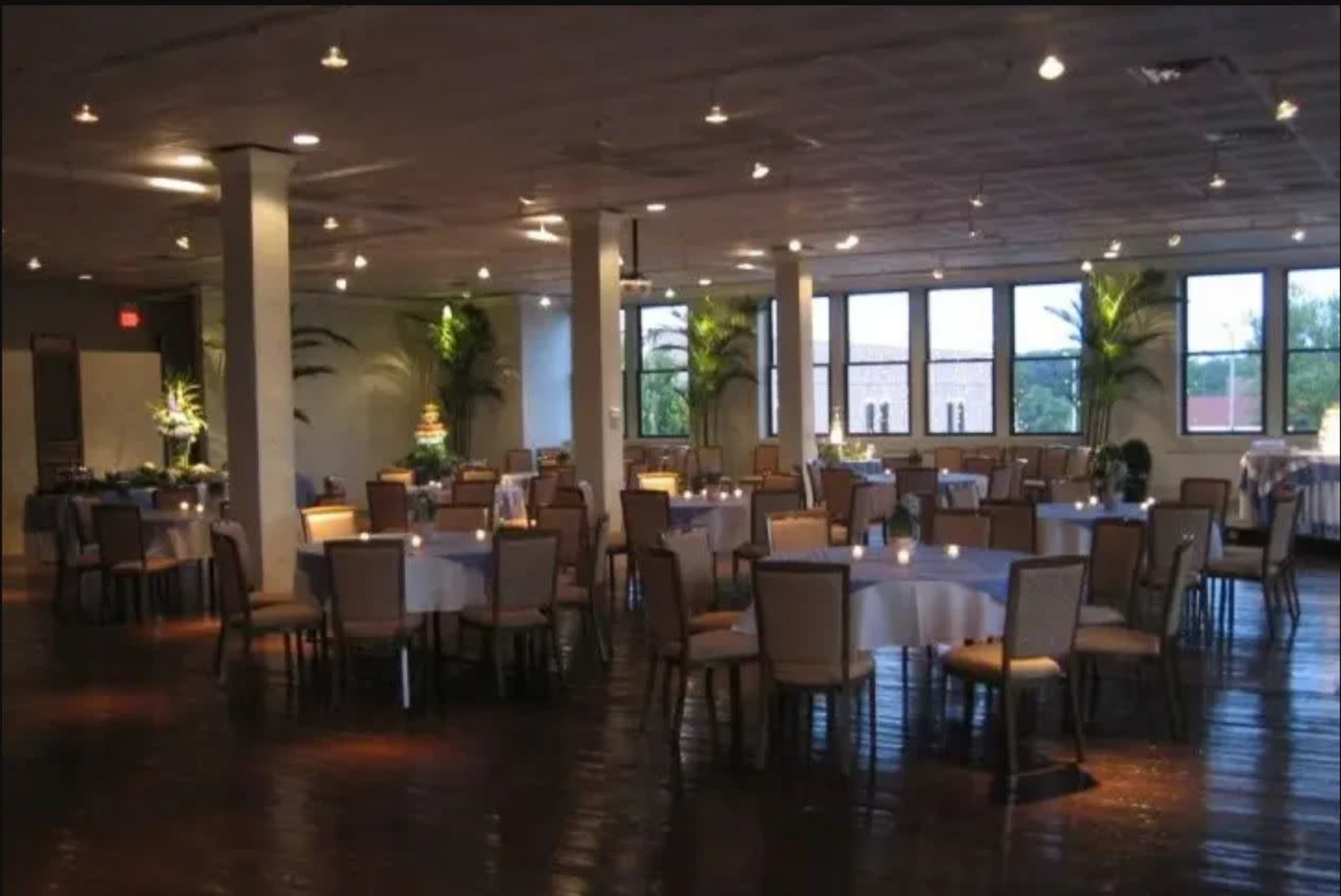 A spacious banquet hall with round tables set for dining, illuminated by warm lighting and featuring large windows with views of greenery outside.