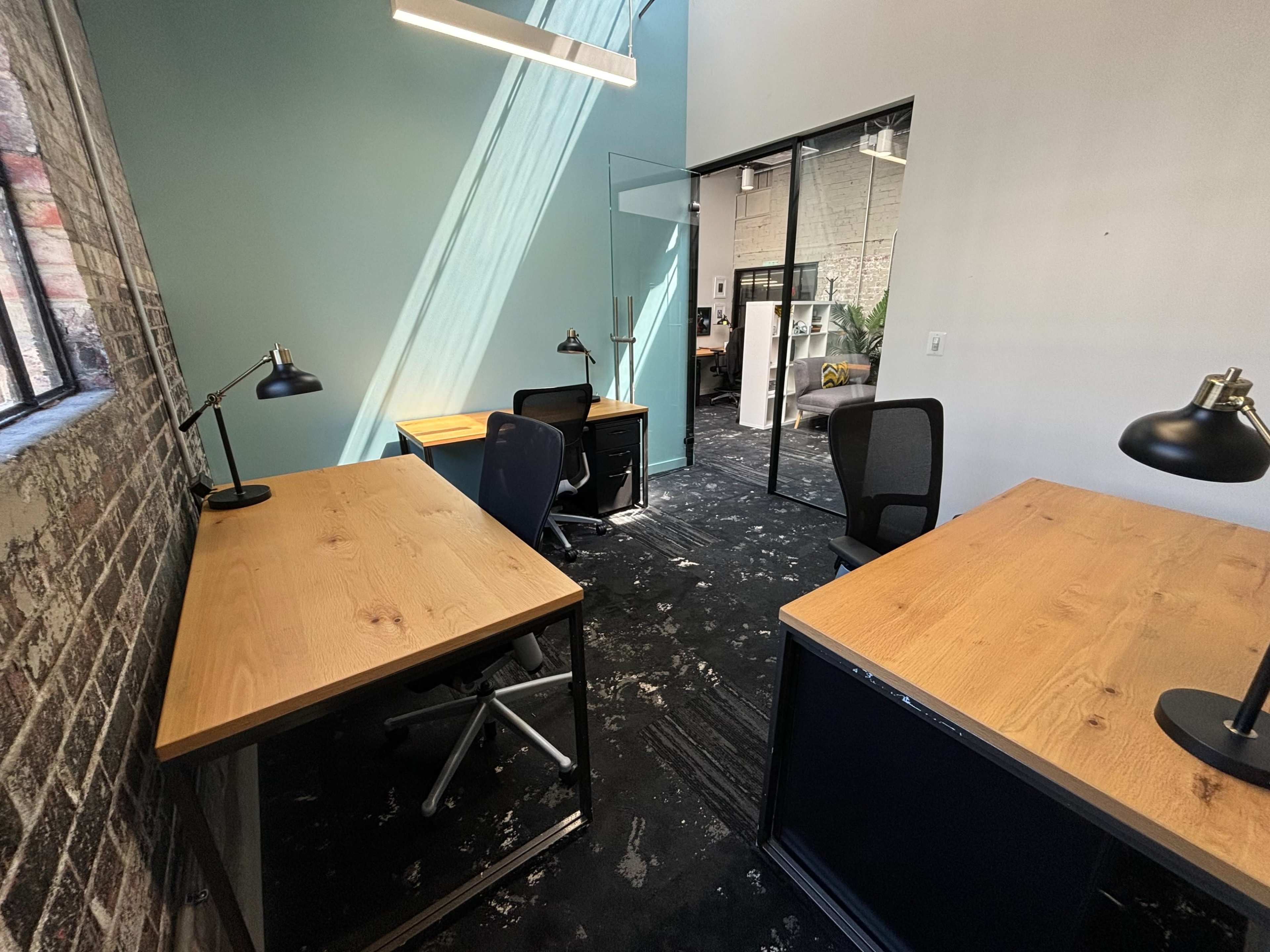 A modern office space with two desks, black office chairs, and large windows casting light onto a textured wall.