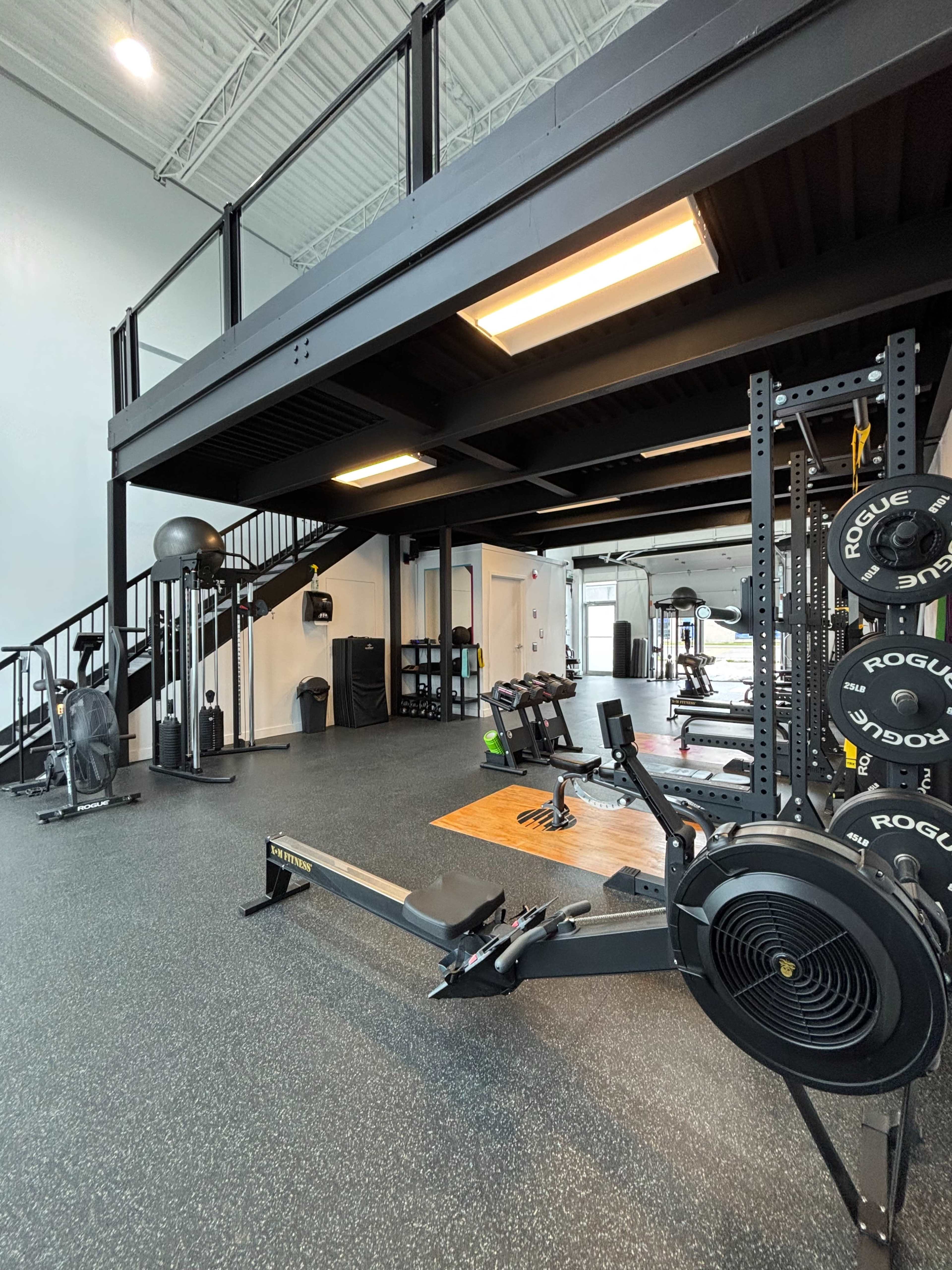 The image shows a spacious gym area featuring various exercise equipment, including rowing machines and weightlifting benches, with a second floor visible in the background.