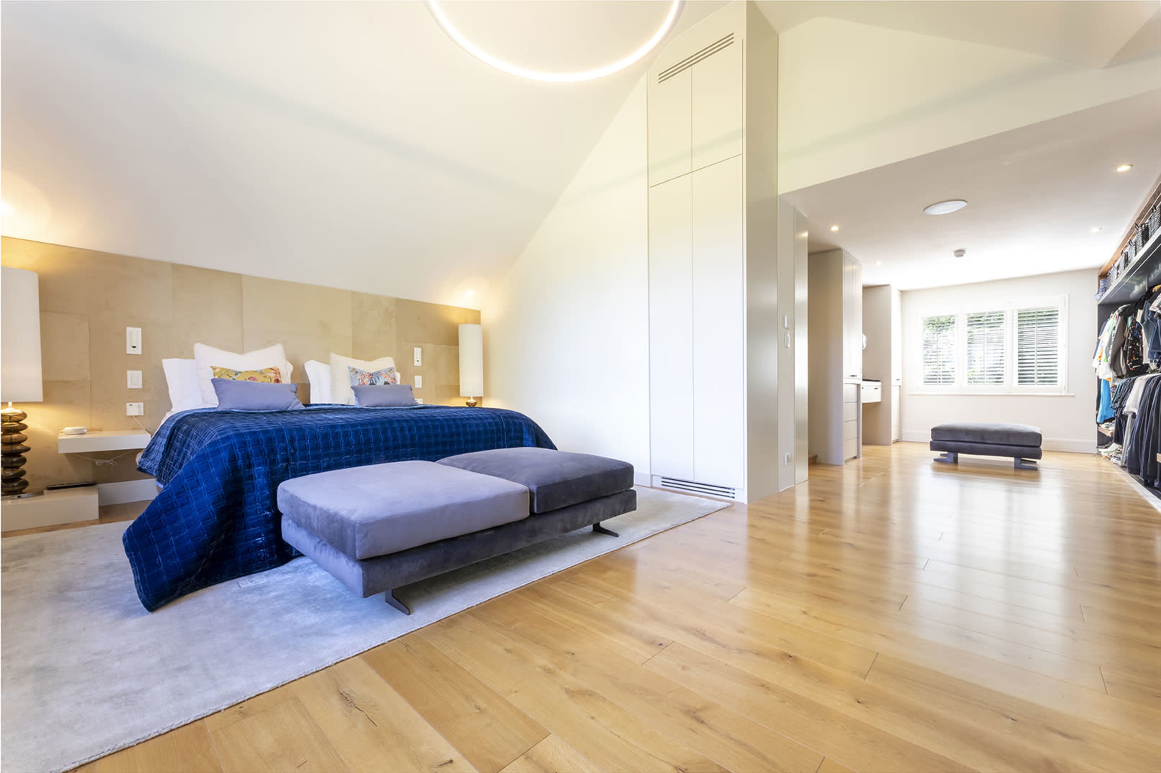 A spacious bedroom features a large bed with a blue blanket, two bedside tables, and a lounge area with a gray ottoman, while a closet is visible on the right side of the room.