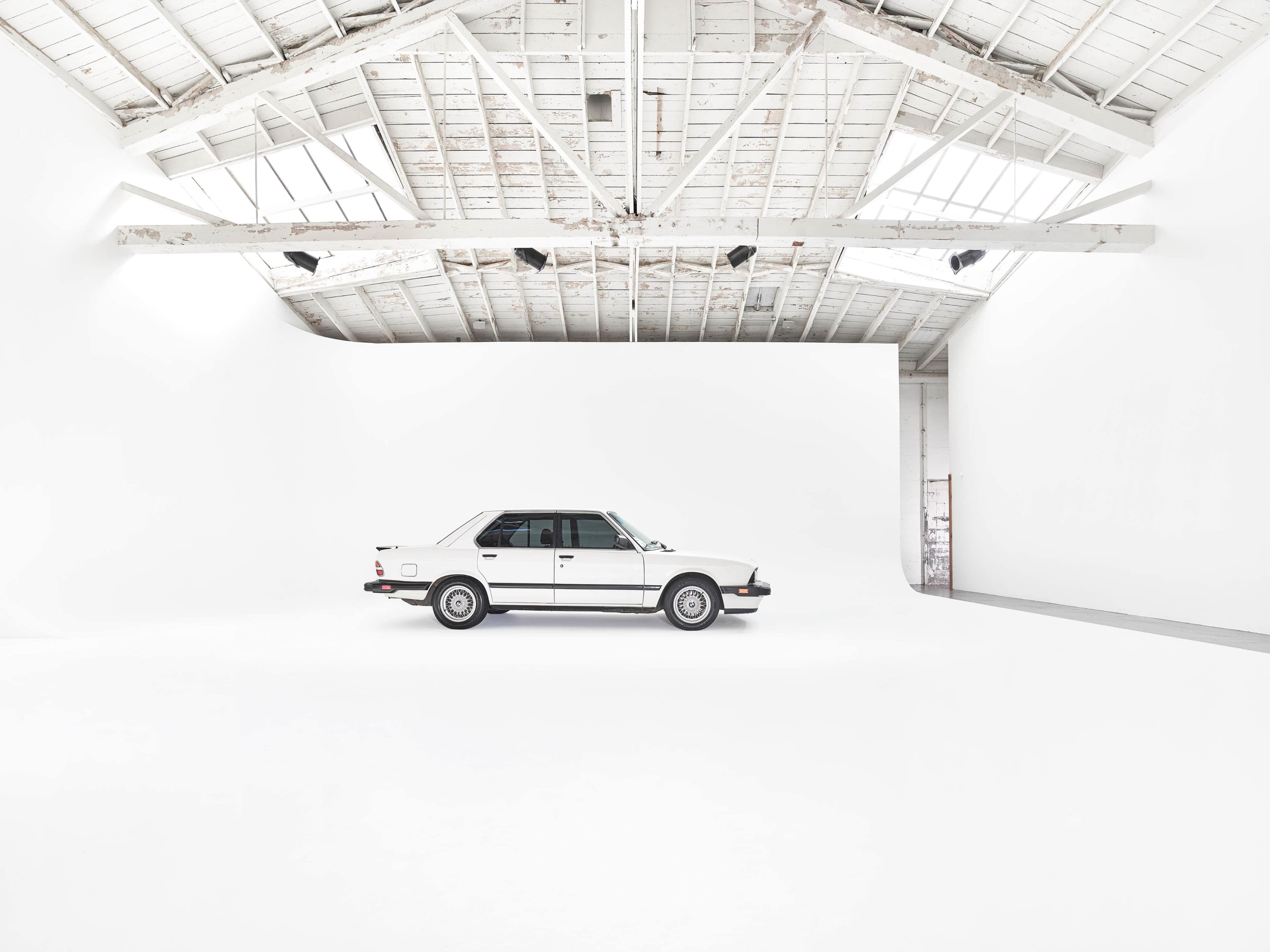 A white car is parked in a spacious, minimalist studio with a high ceiling and white walls.