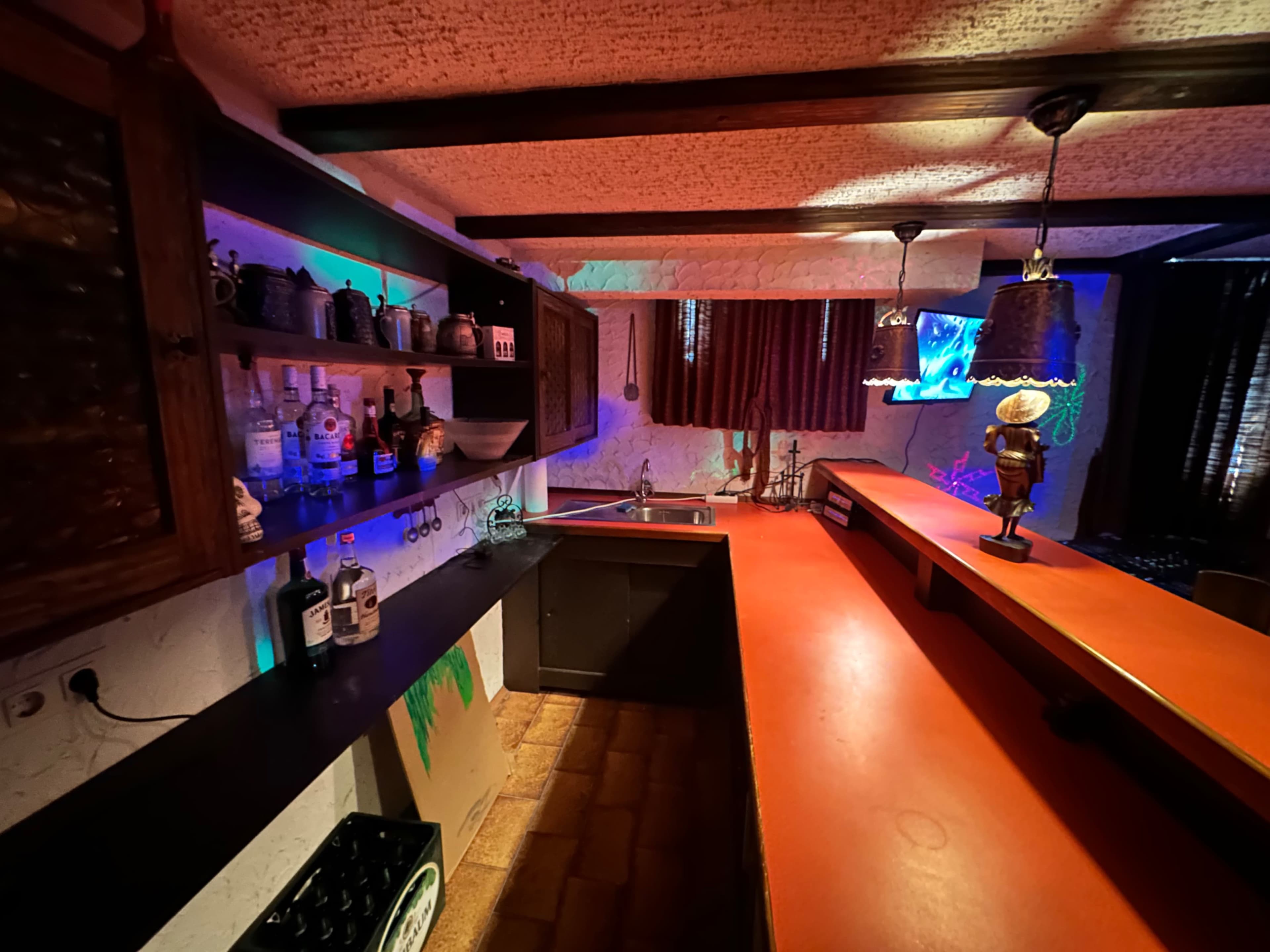 A dimly lit kitchen with a long red countertop, shelves holding various bottles, and colorful ambient lighting.