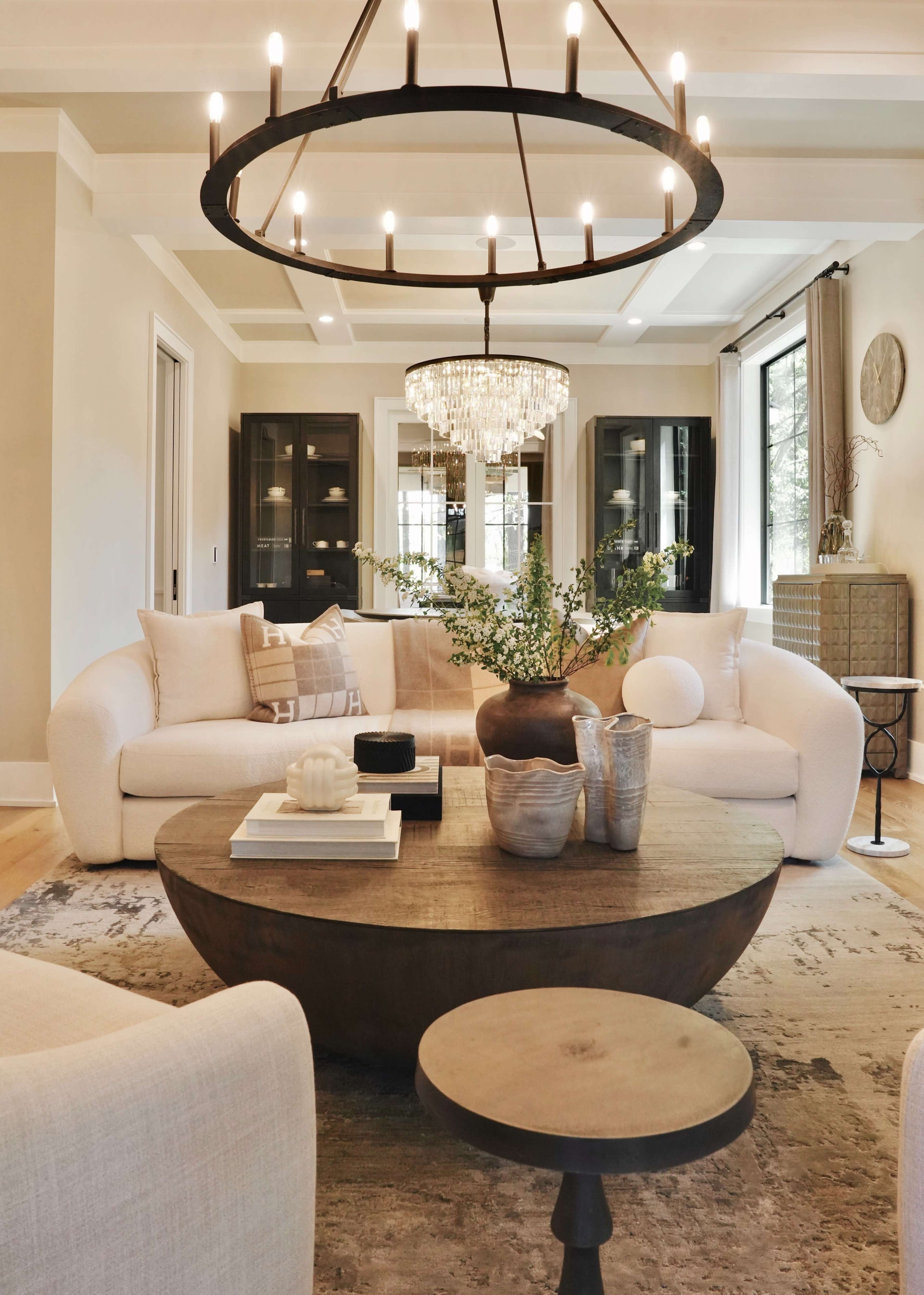 A modern living room features a large round wooden coffee table, a white sectional sofa, and a striking chandelier above, with decor items arranged on the table and shelves in the background.