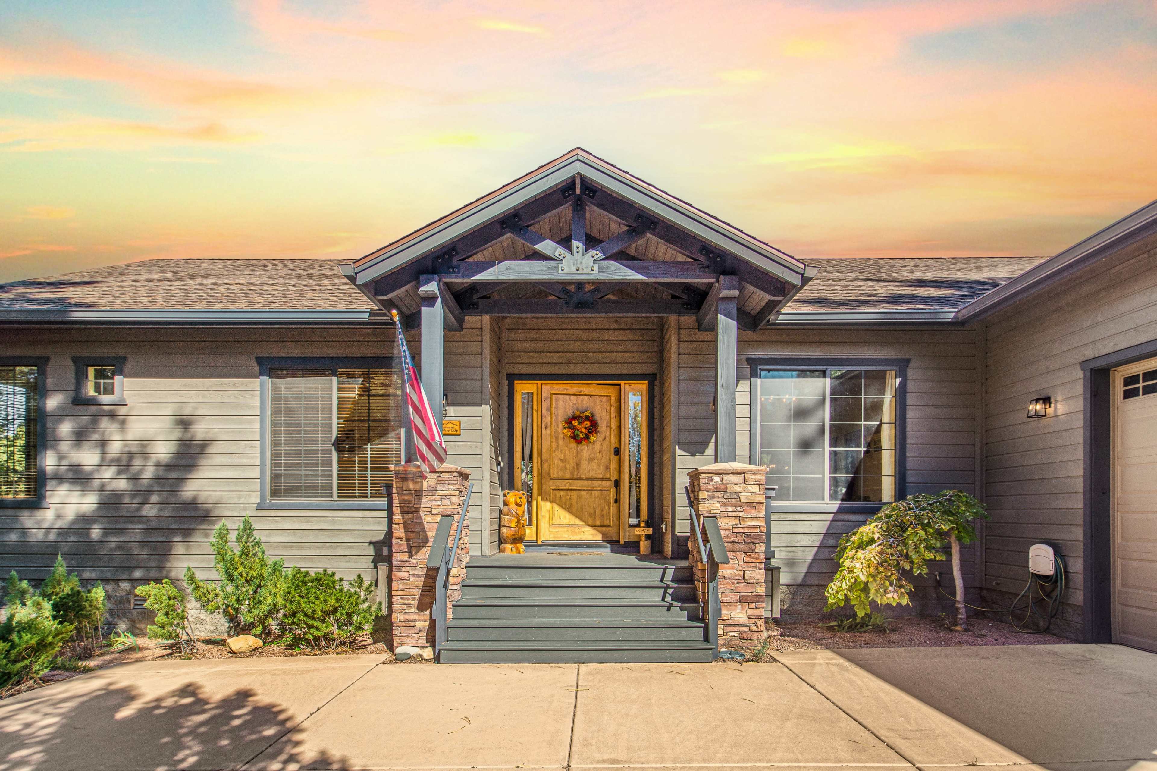 A single-story house with a front porch, wooden door, American flag, and a decorative wreath, framed by stone accents and surrounding greenery.