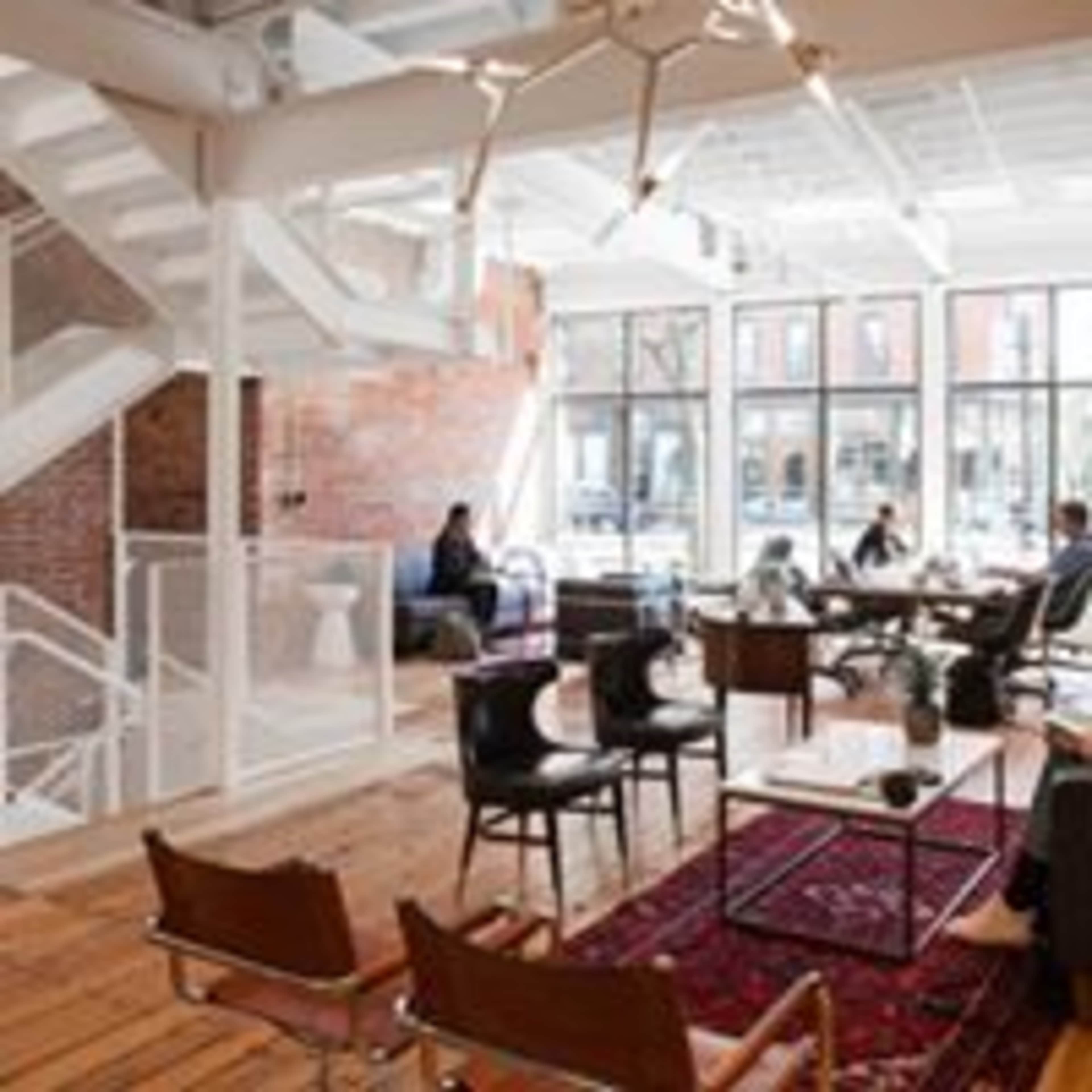 The image shows a modern open-plan lounge area with wooden flooring, large windows, and a mix of seating arrangements including chairs and sofas.