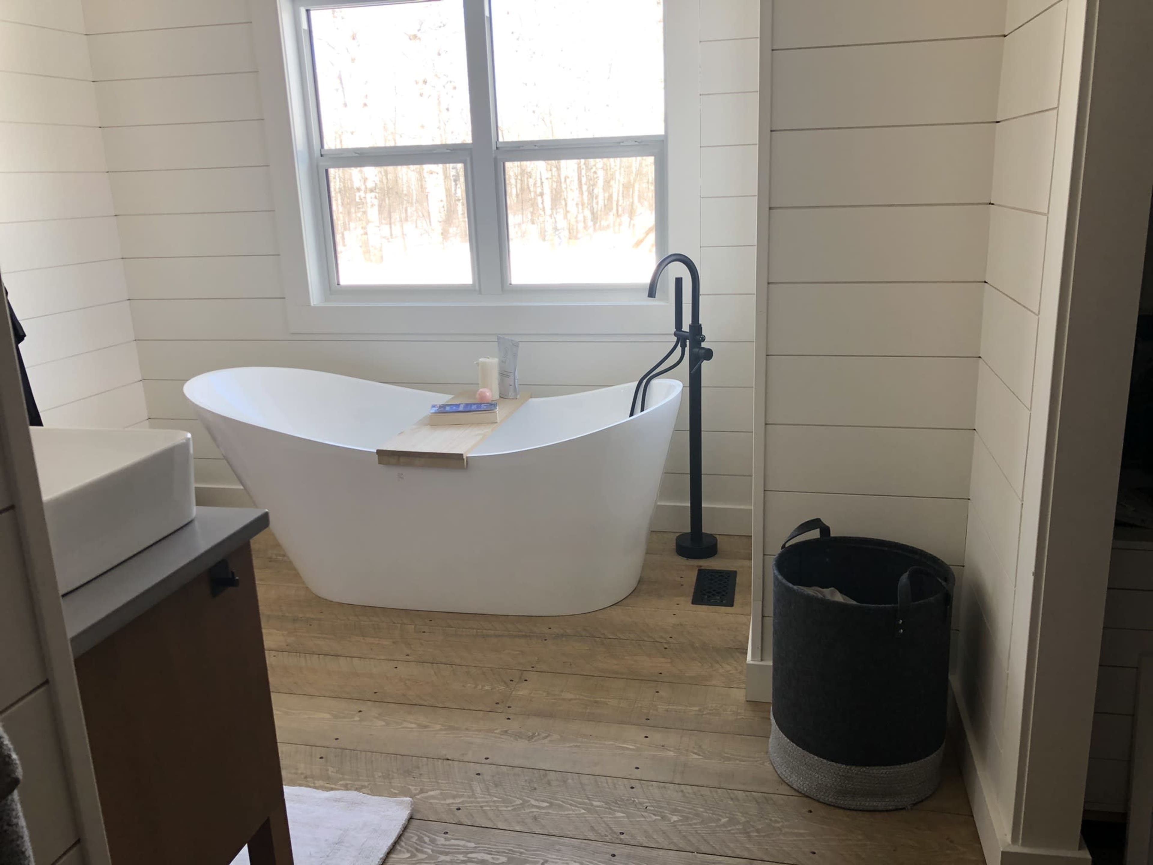 A modern bathroom featuring a freestanding white bathtub next to a large window, set on wooden flooring with a gray basket nearby.