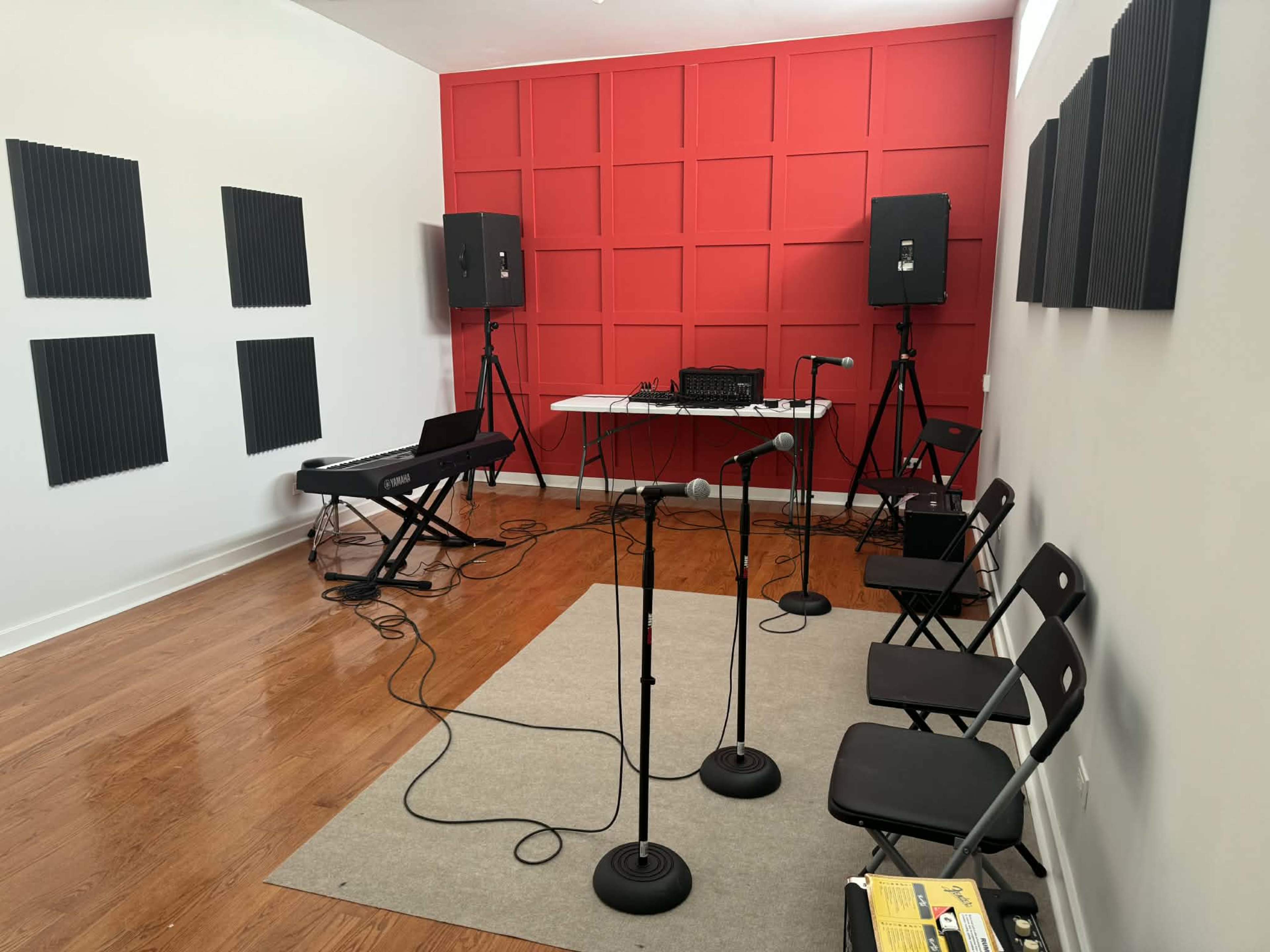 The image shows a music rehearsal space with a red accent wall, a keyboard on a stand, two speakers on tripods, and several microphones arranged near a table.