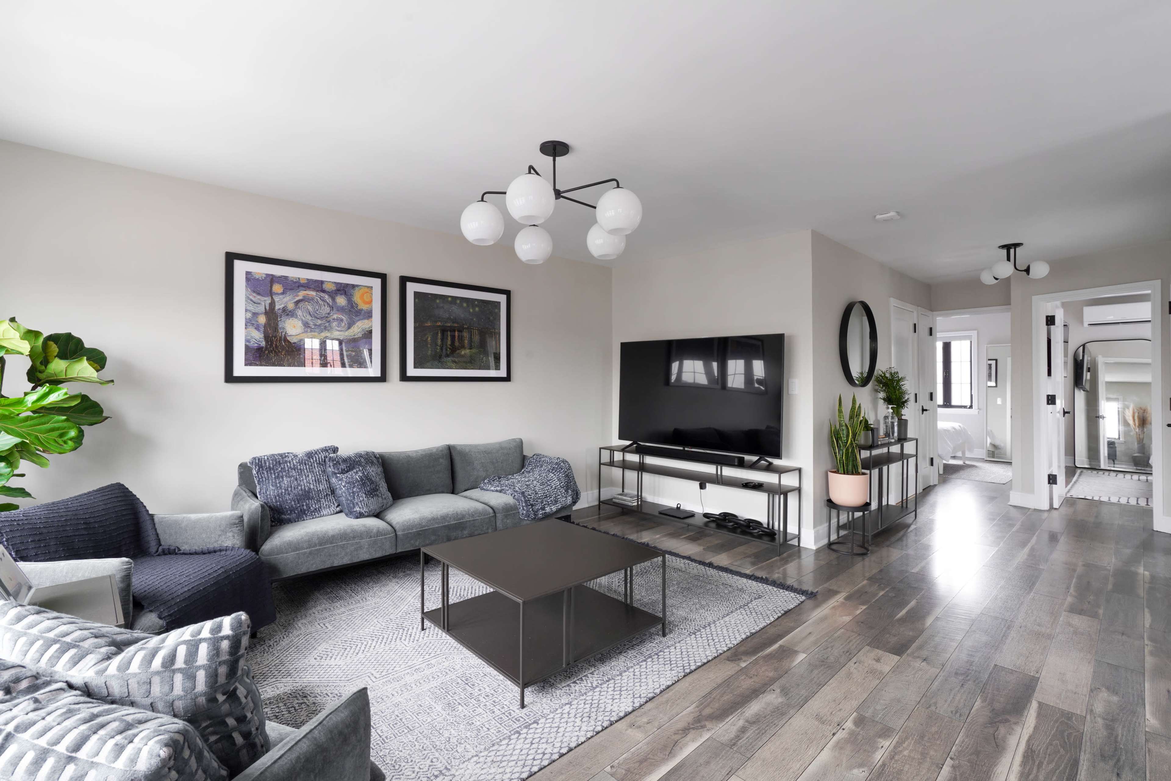 The image shows a modern living room with a dark gray sofa, a coffee table, and a large television on a white stand, featuring artwork on the walls and a plant in the corner.