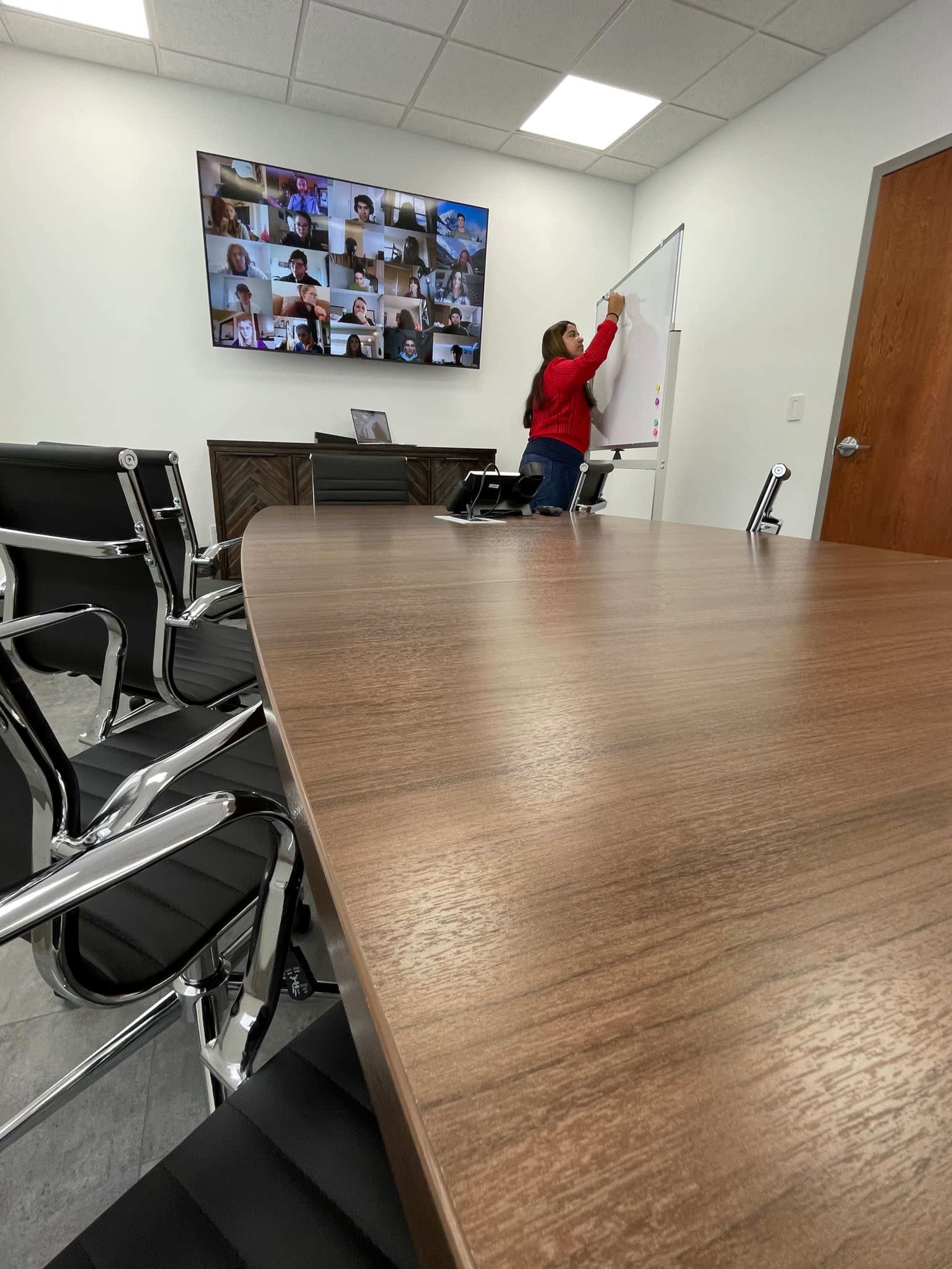 A person writes on a whiteboard in a conference room equipped with a large screen displaying a video call with multiple participants.