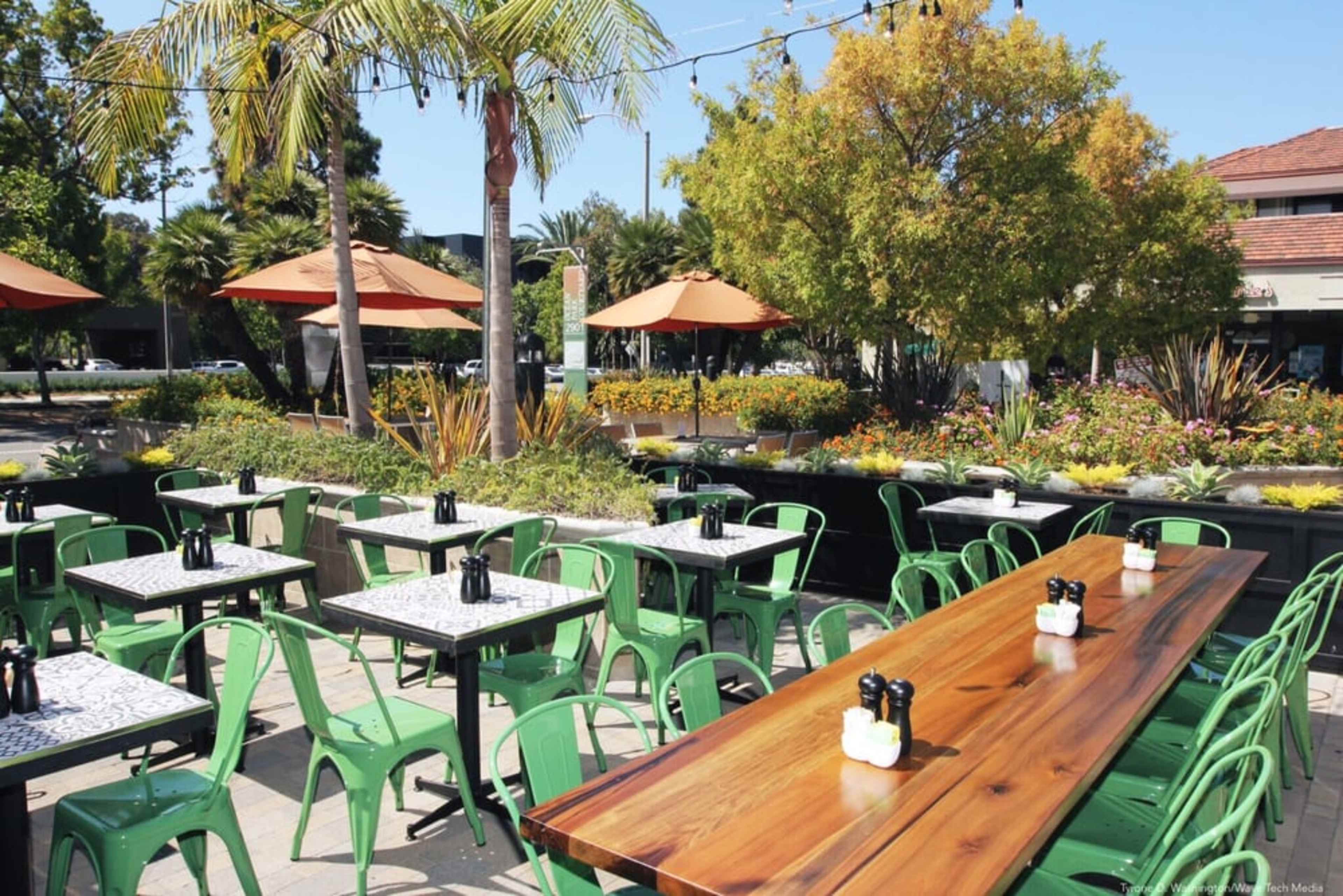 A patio area features green metal chairs and wooden tables set up under orange umbrellas surrounded by plants and flowers.