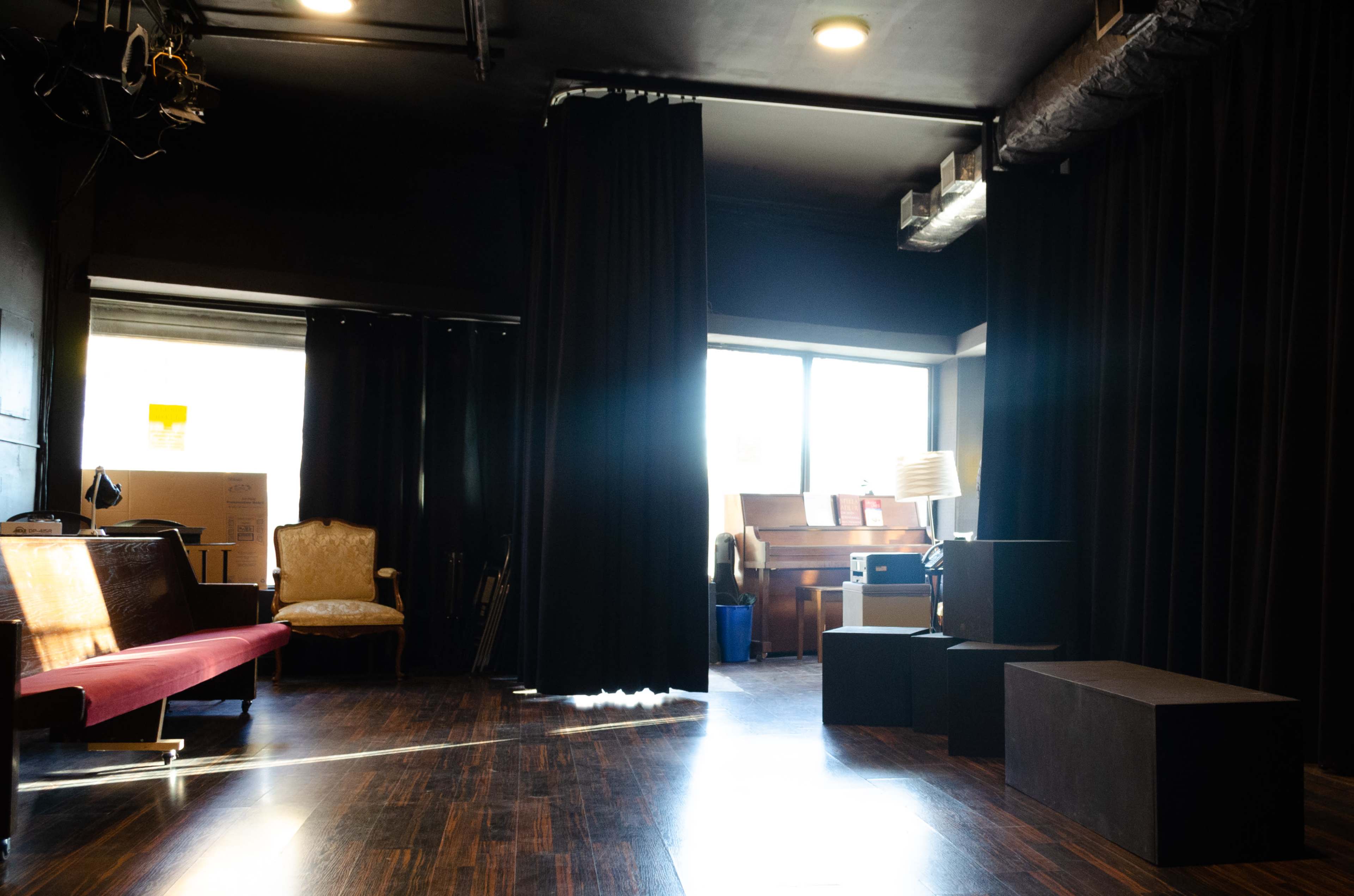 A small performance space features a red bench, black curtains, a piano, and an armchair, with large windows allowing natural light.