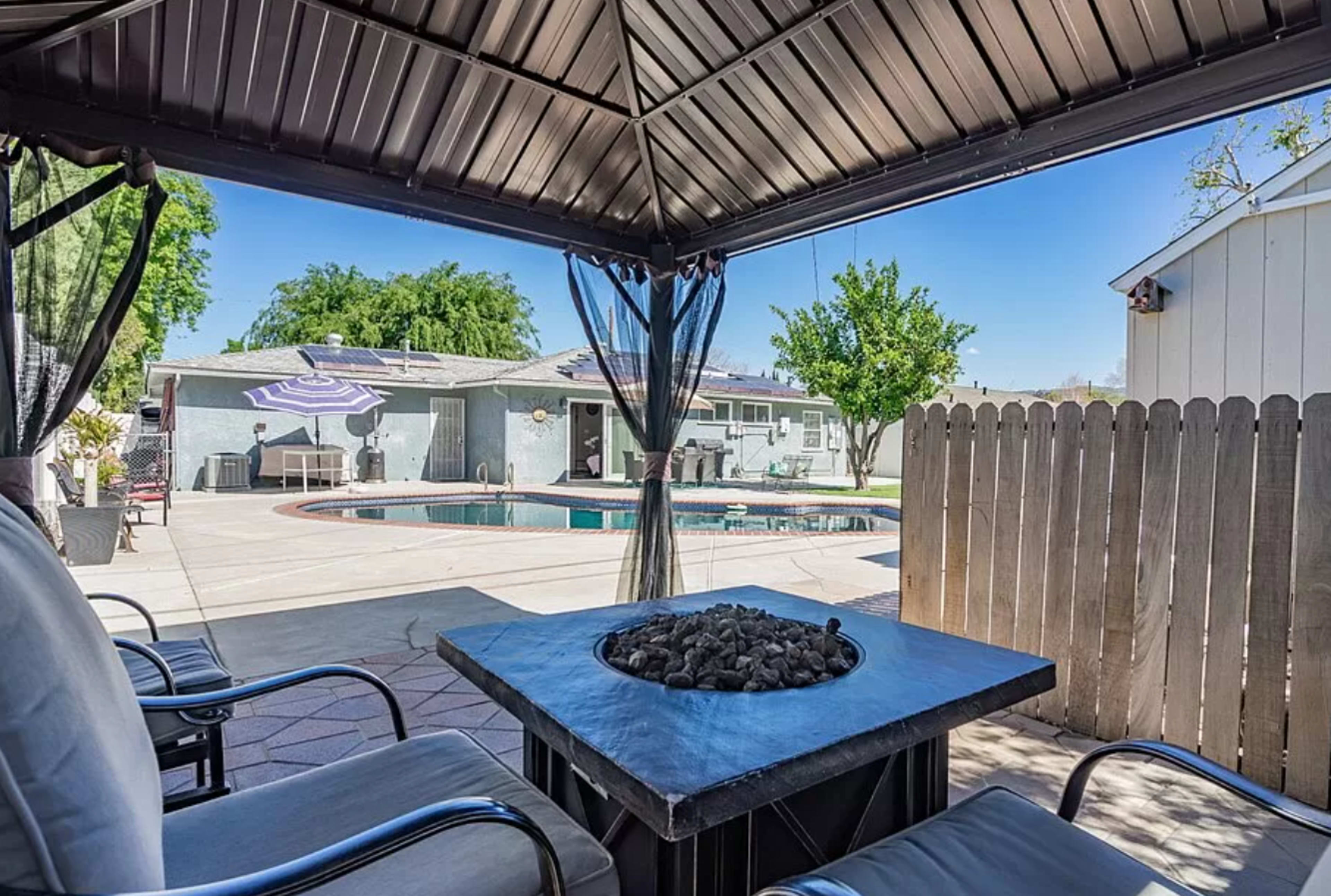The image shows a backyard with a pool, a gazebo, and seating around a fire pit, enclosed by a wooden fence.