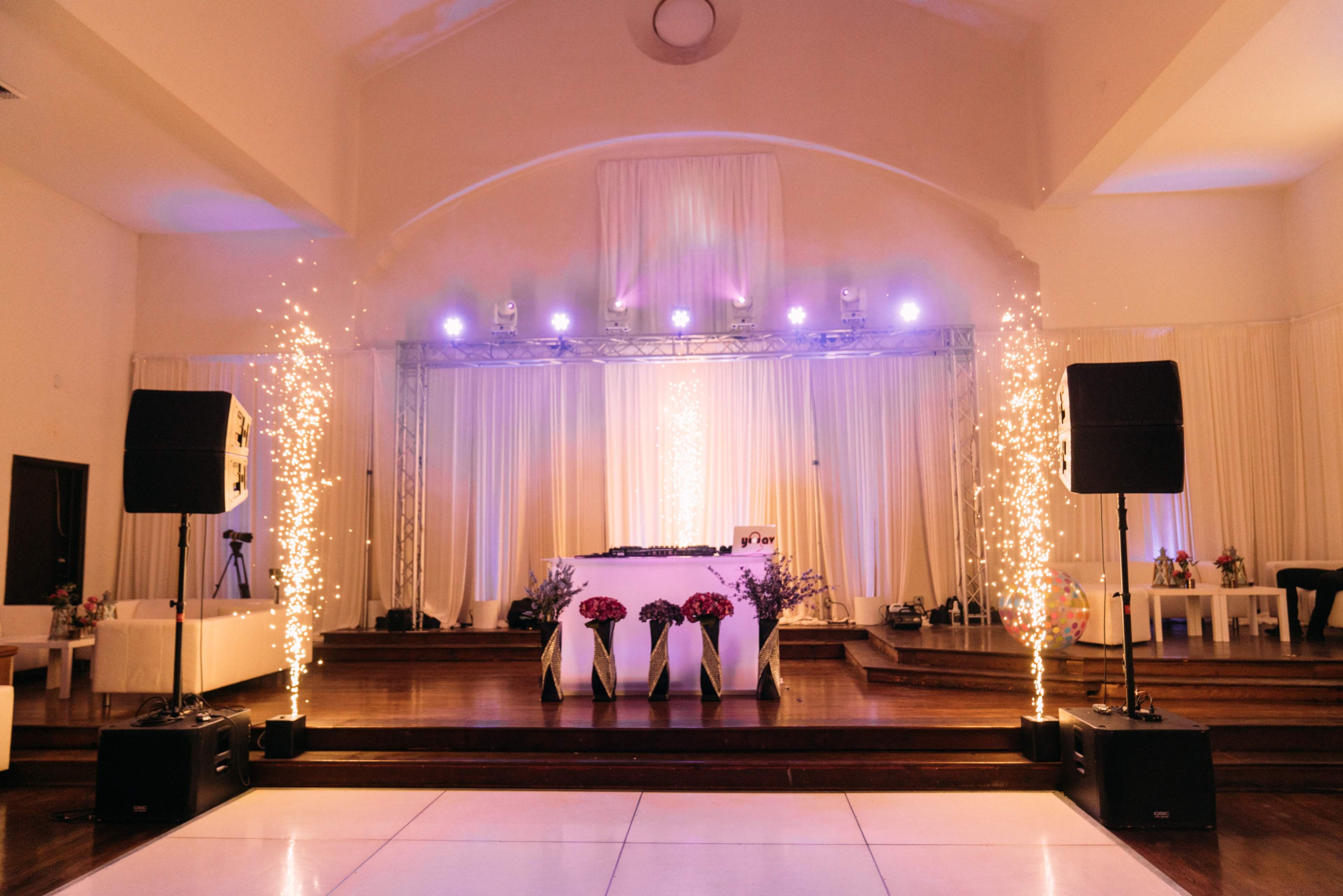 The scene features a decorated event space with a DJ booth, soft lighting, and floral arrangements on stage, flanked by two tall speakers and decorative sparkler effects.