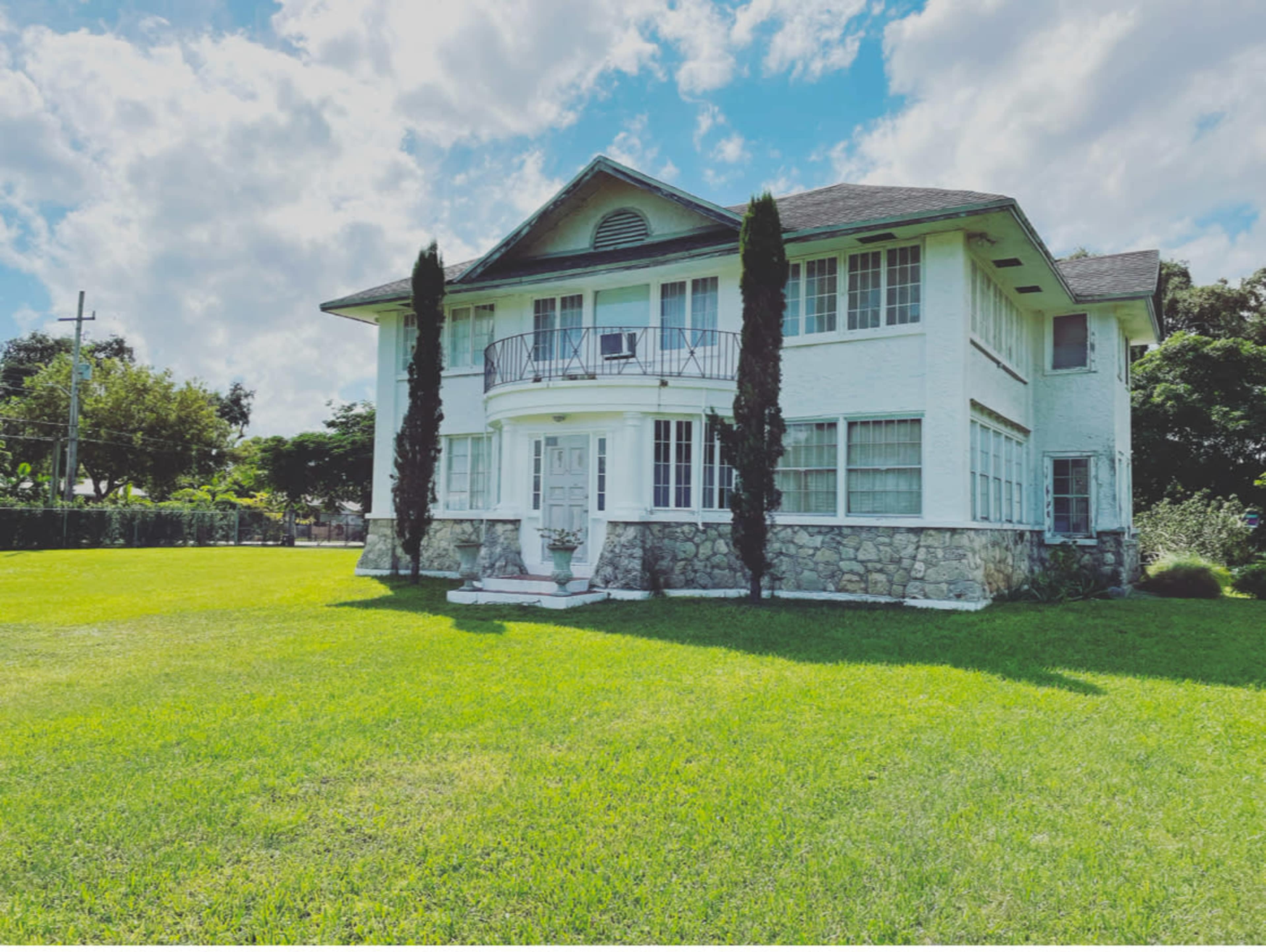 A large, two-story white house with a stone foundation and manicured lawn is set against a bright, cloudy sky.