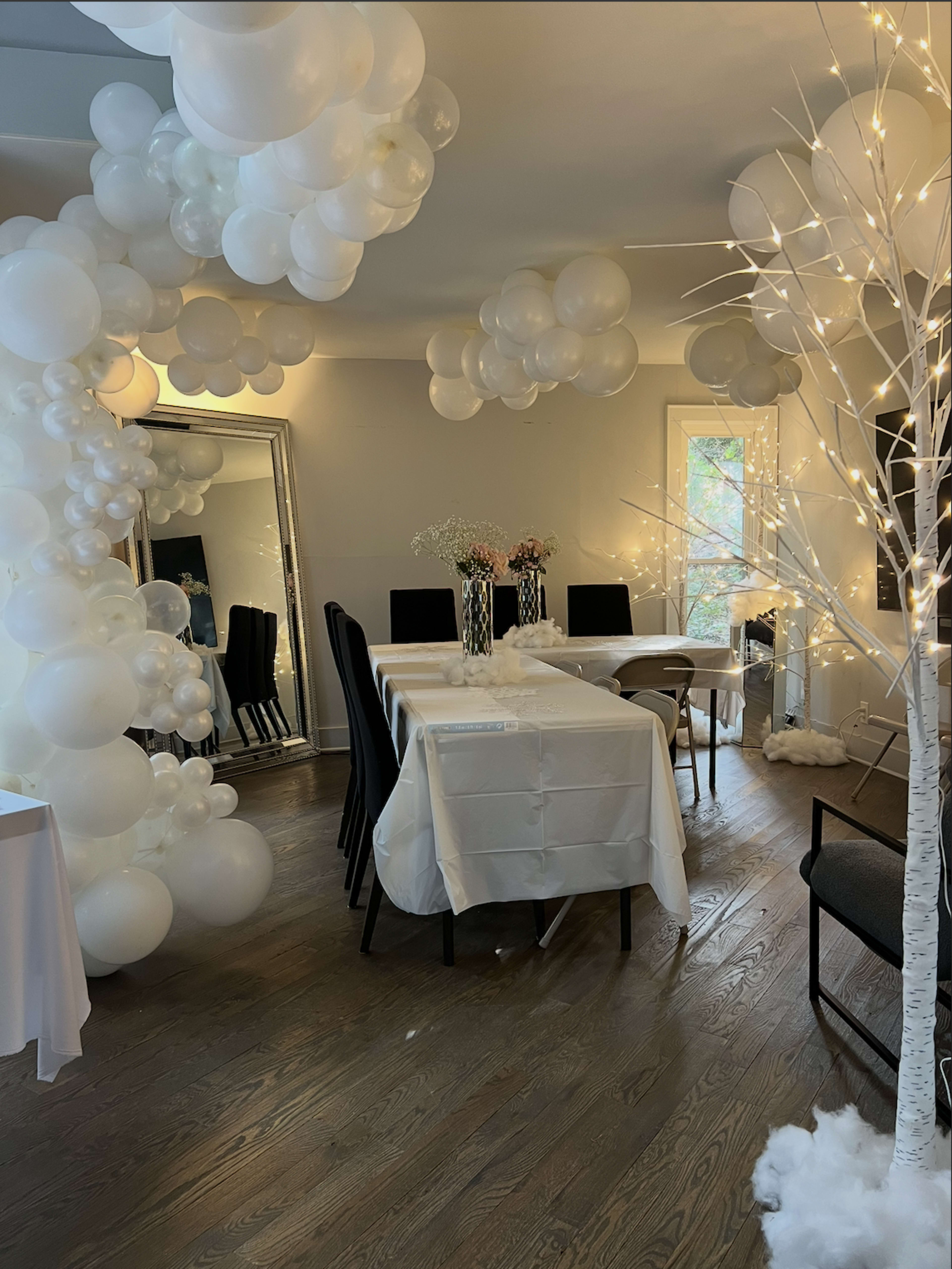 A dining room is decorated with white balloons and artificial trees adorned with lights, featuring a large table set with a white tablecloth and flowers.